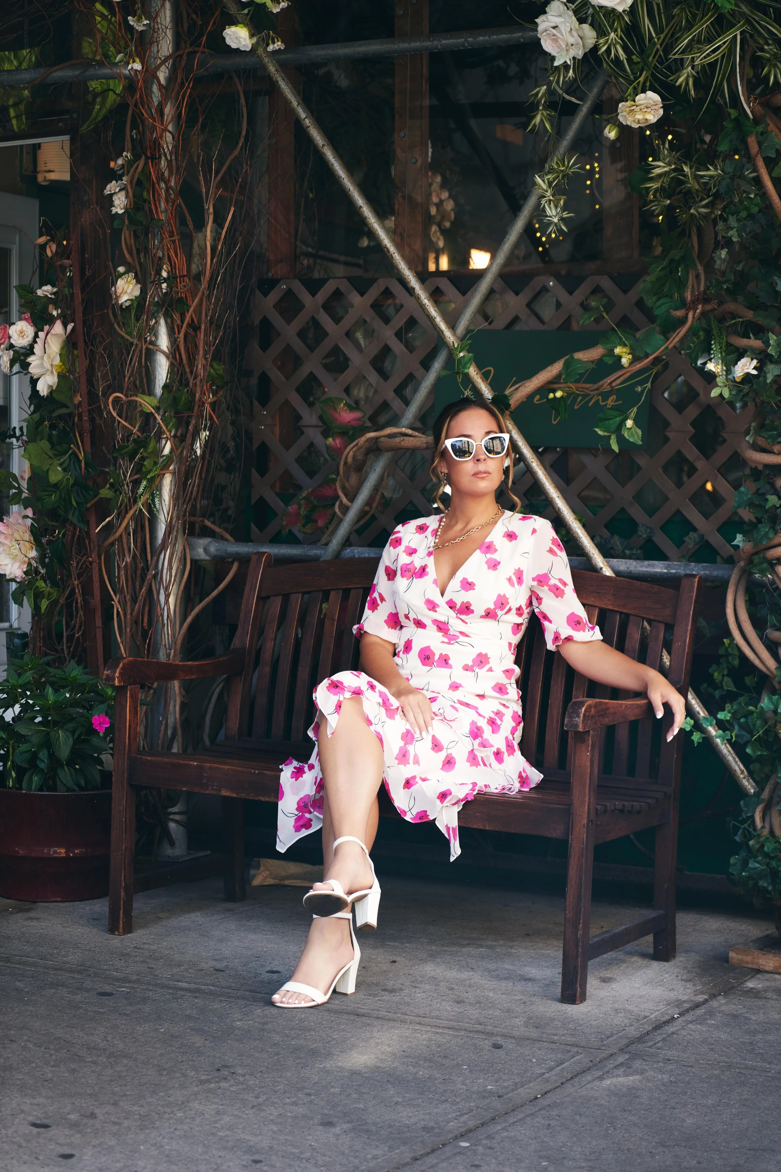A woman sitting on a wooden bench outdoors, wearing a white dress with pink floral pattern, white high-heeled sandals, white sunglasses, and a gold necklace, surrounded by plants and greenery.