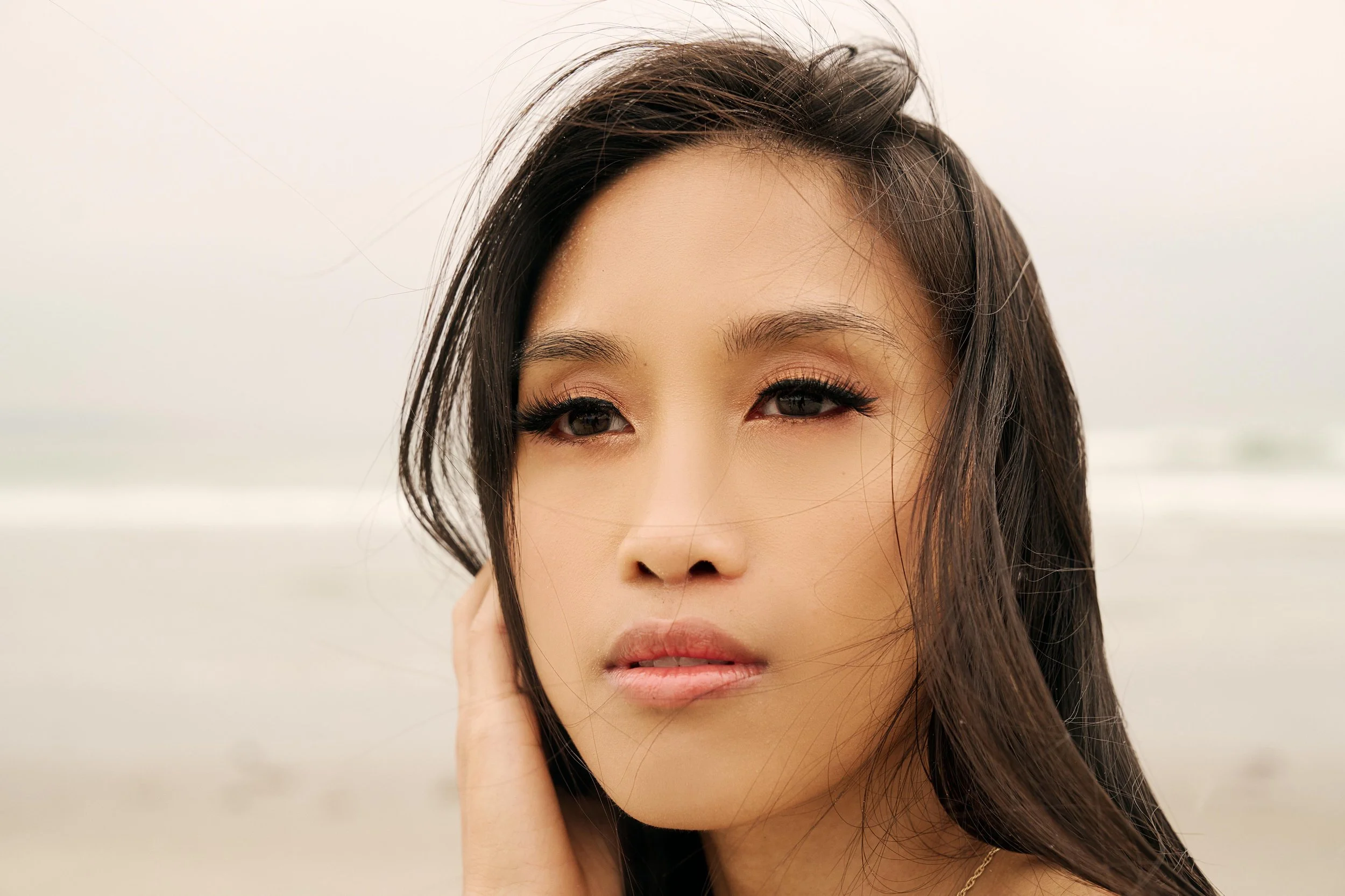 Close-up of a young Asian woman with long dark hair at the beach, looking into the camera with wind-blown hair and a calm expression.