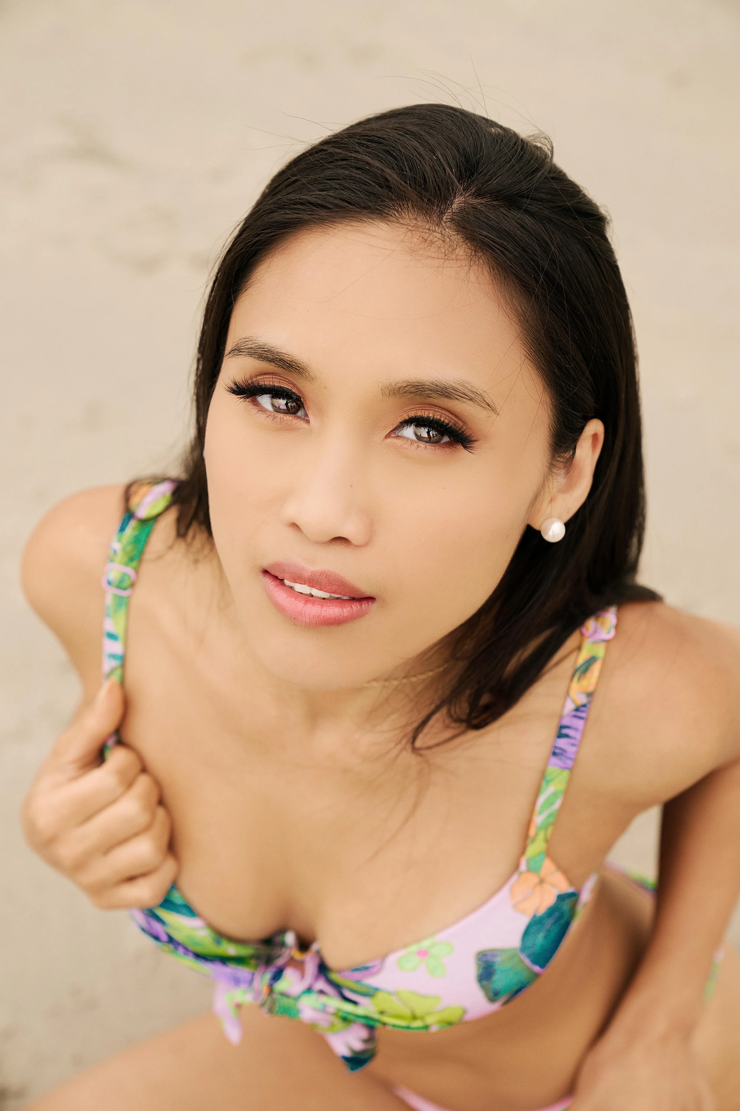 A woman with dark hair and pearl earrings looking up at the camera, wearing a colorful floral bikini on the beach.