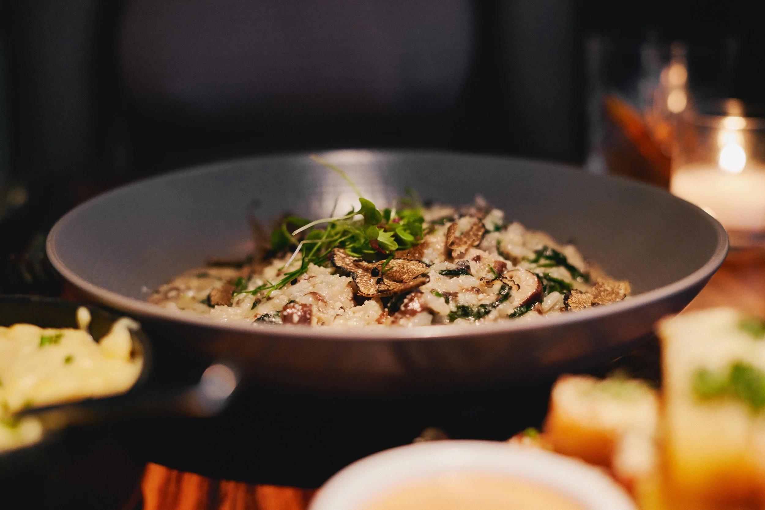 A bowl of creamy pasta with mushrooms, greens, and garnished with microgreens, on a table with a lit candle beside it.