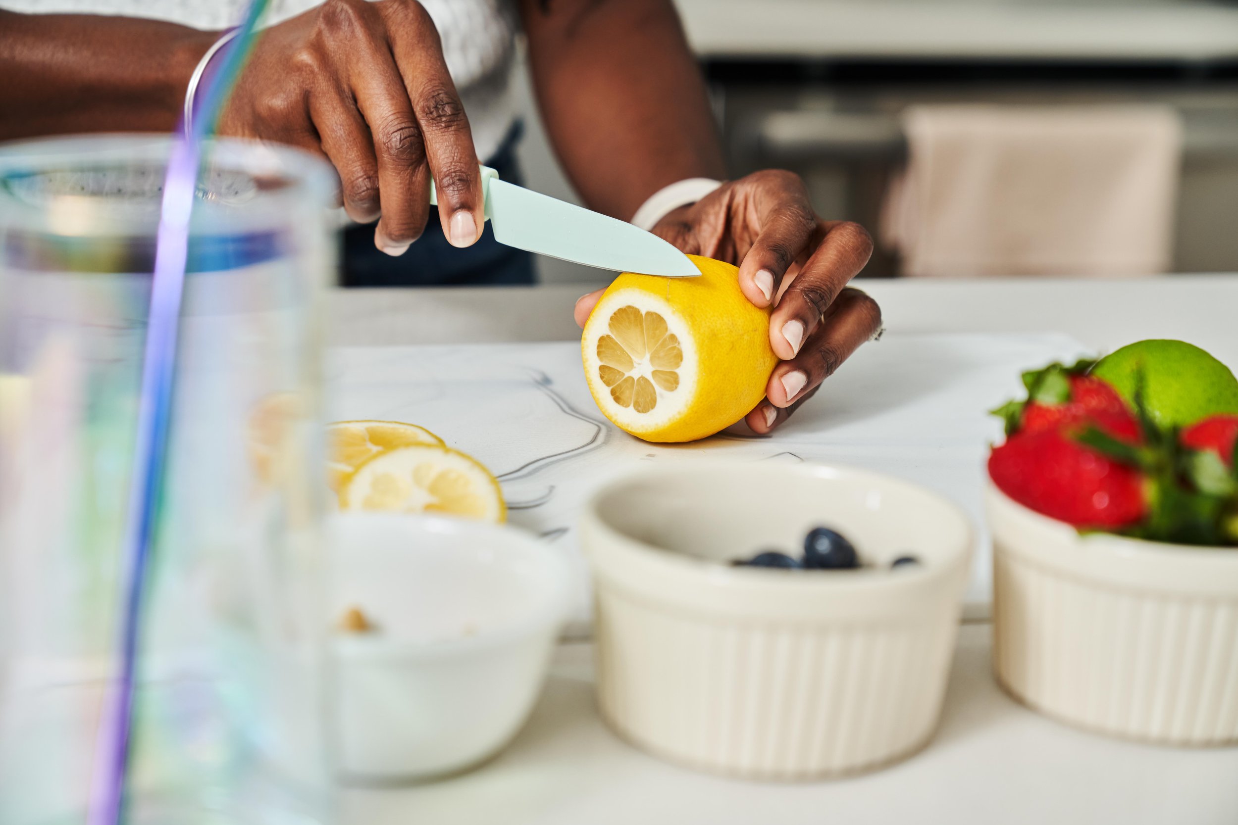 A person slices a lemon with a knife on a white kitchen counter. There are bowls of strawberries, blueberries, lemon slices, and a lemon in the background.