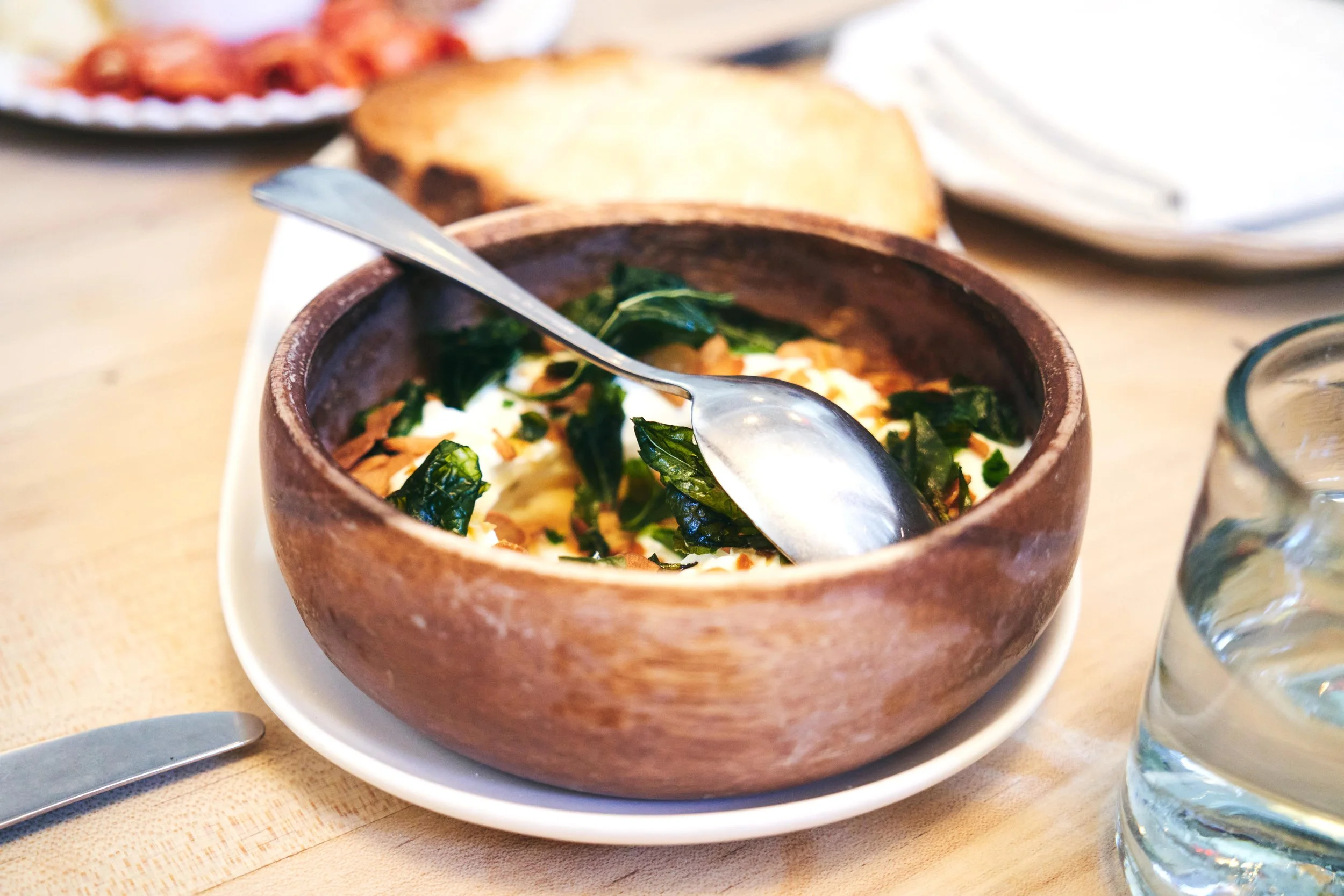 A bowl of spinach and feta cheese salad with toasted almonds, with bread in the background on a wooden table. 