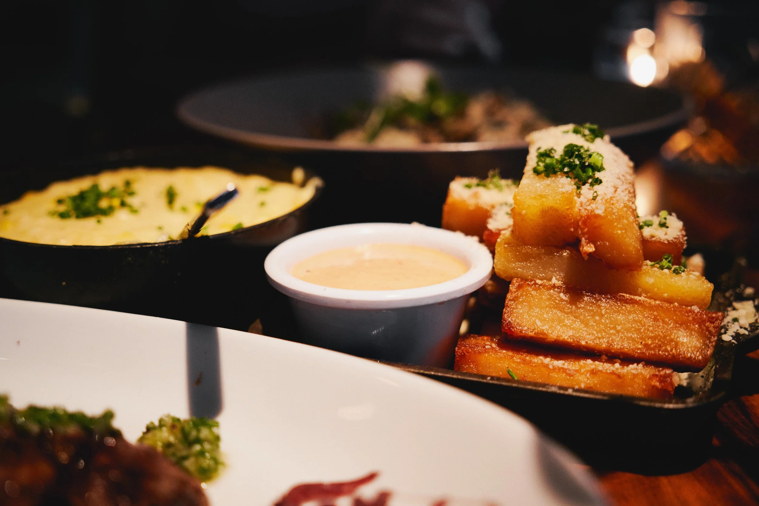 Fried pork belly slices topped with chopped green onions, served with a side of dipping sauce and a small dish of mashed potatoes garnished with green herbs, in a dimly lit restaurant setting.