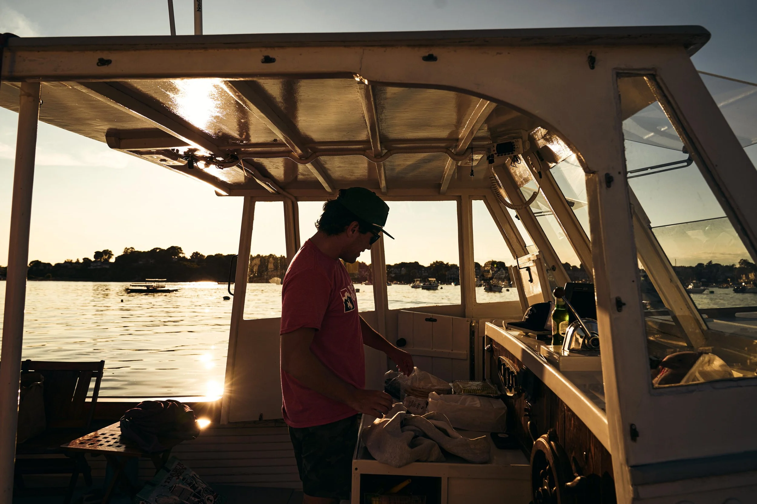 A man in a red shirt and cap preparing food inside a boat during sunset, with water and trees in the background.