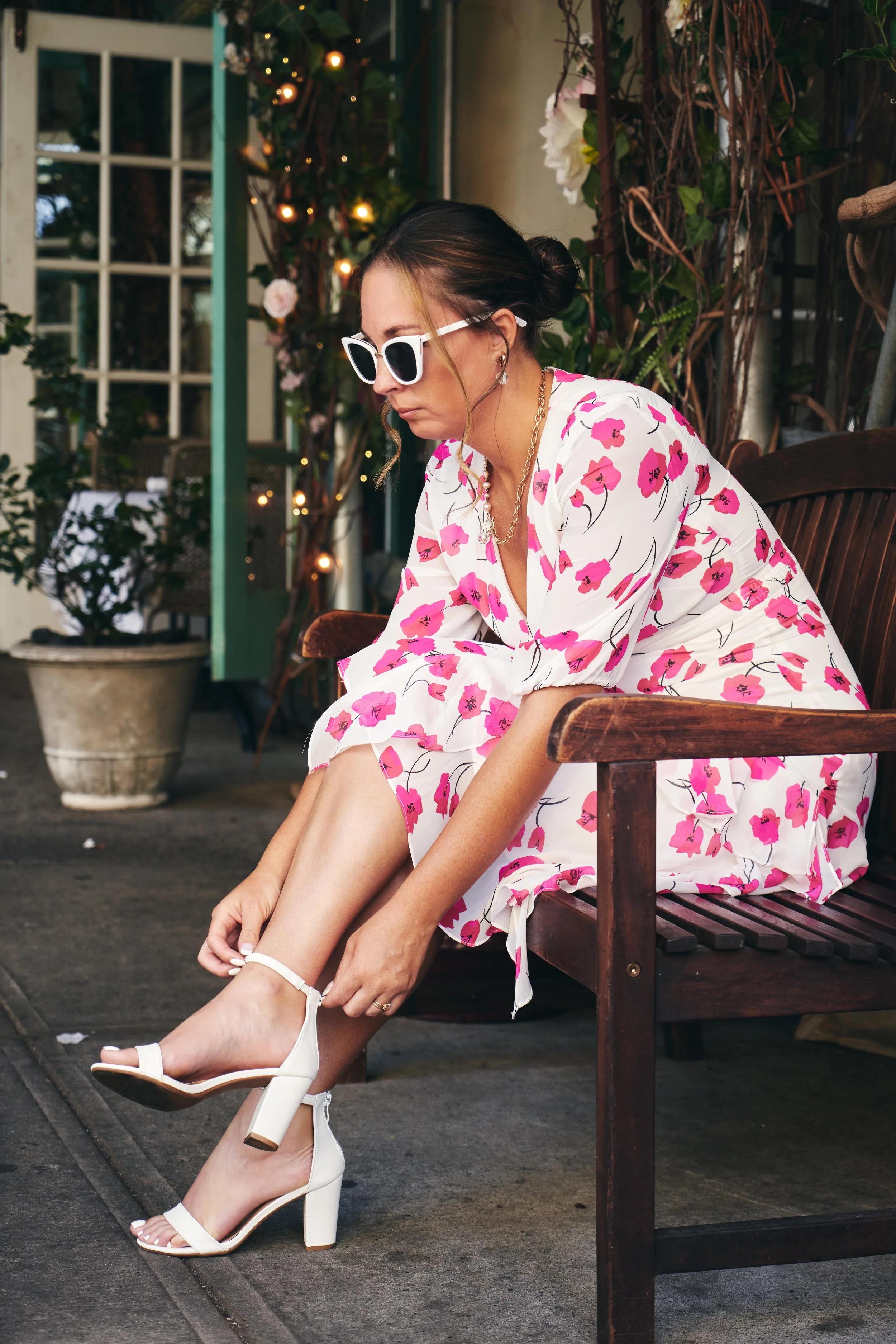 A woman in a white dress with pink floral prints, wearing white high-heeled sandals, sitting on a wooden bench, adjusting her shoe strap, with plants and fairy lights in the background.