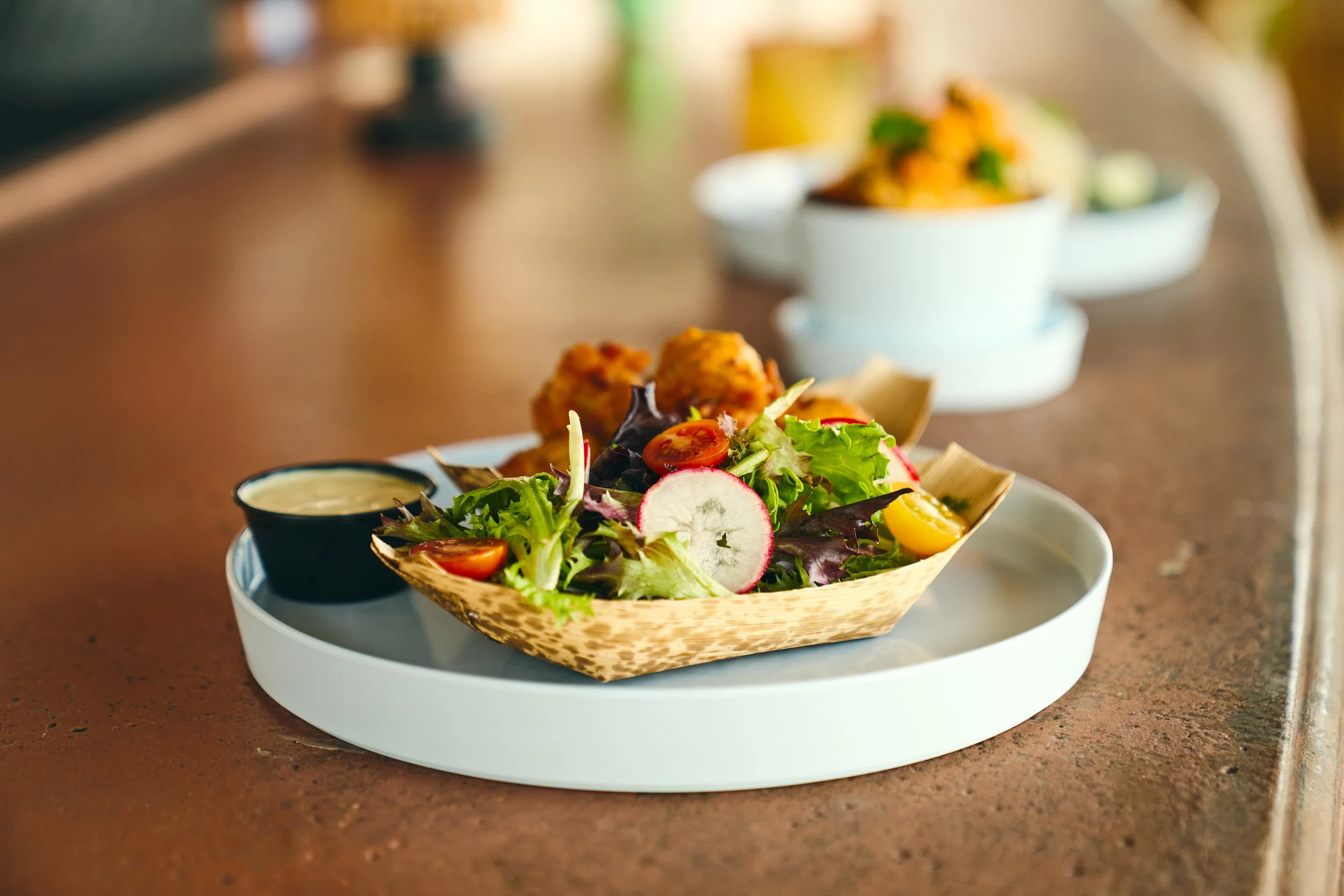 A plate of fresh salad with cherry tomatoes, radish slices, mixed greens, and two fried appetizers beside a small container of dressing, on a white tray in a restaurant setting.