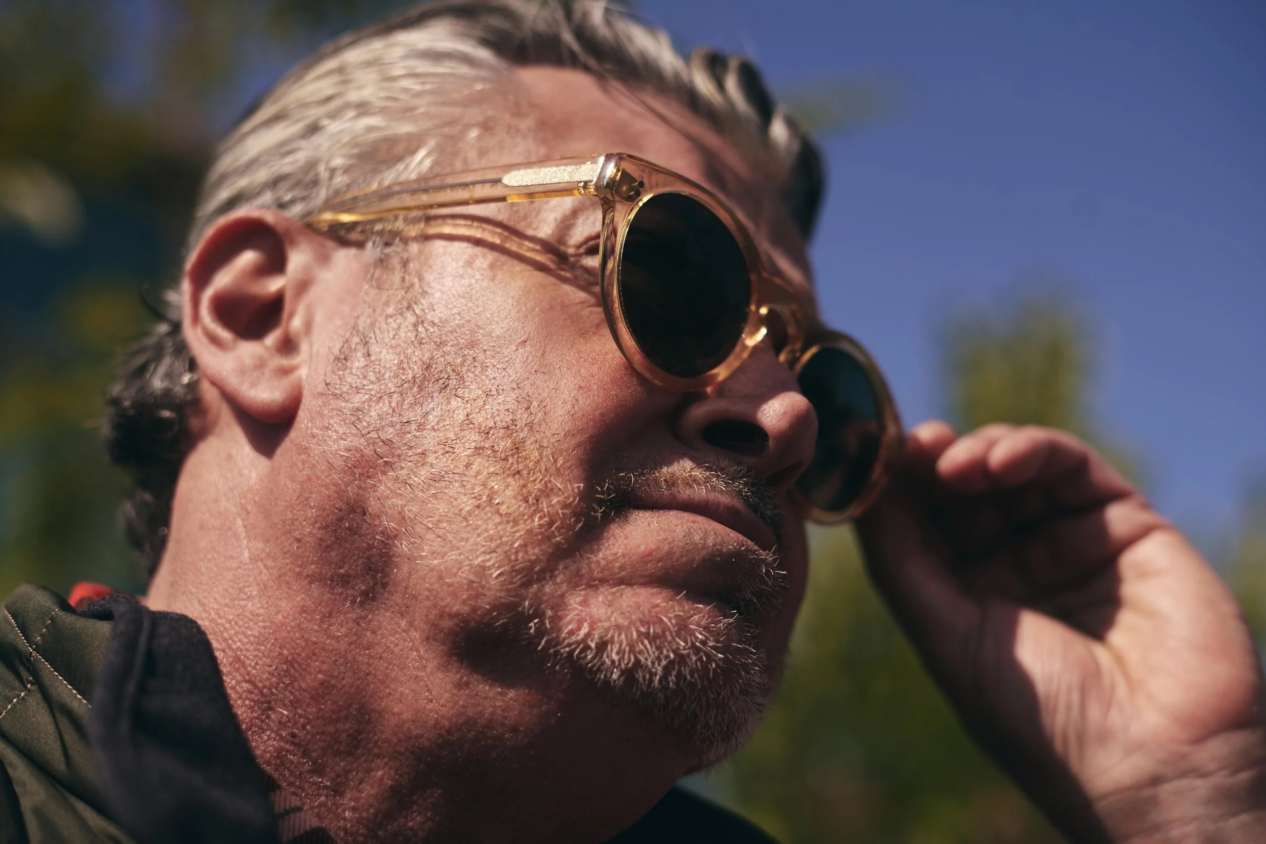 Close-up of an older man with gray hair and beard, wearing round sunglasses with a gold frame, adjusting his sunglasses with his hand, outdoors on a sunny day.
