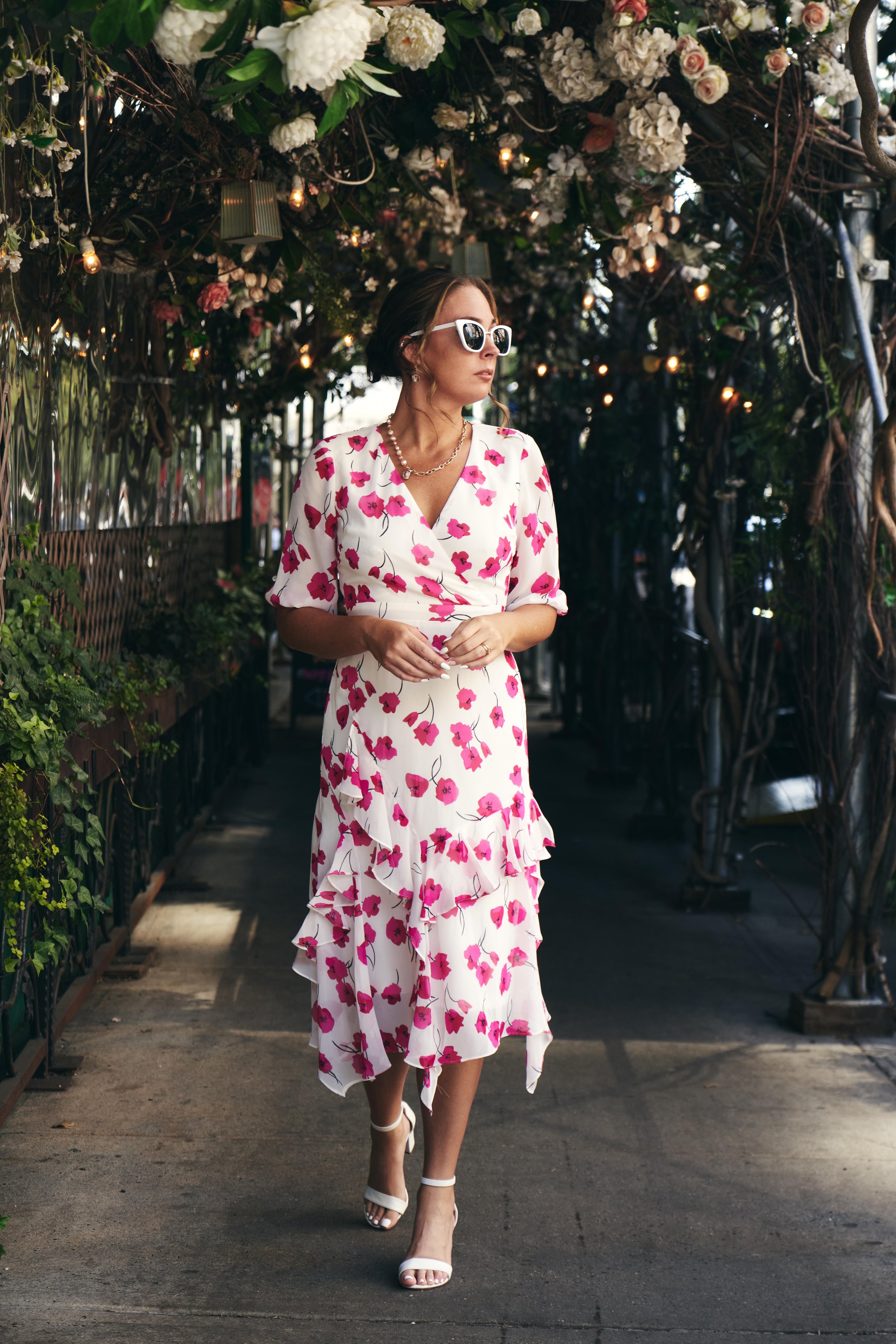 A woman walking through a floral archway, wearing a white dress with pink flowers, white high heels, sunglasses, and jewelry.