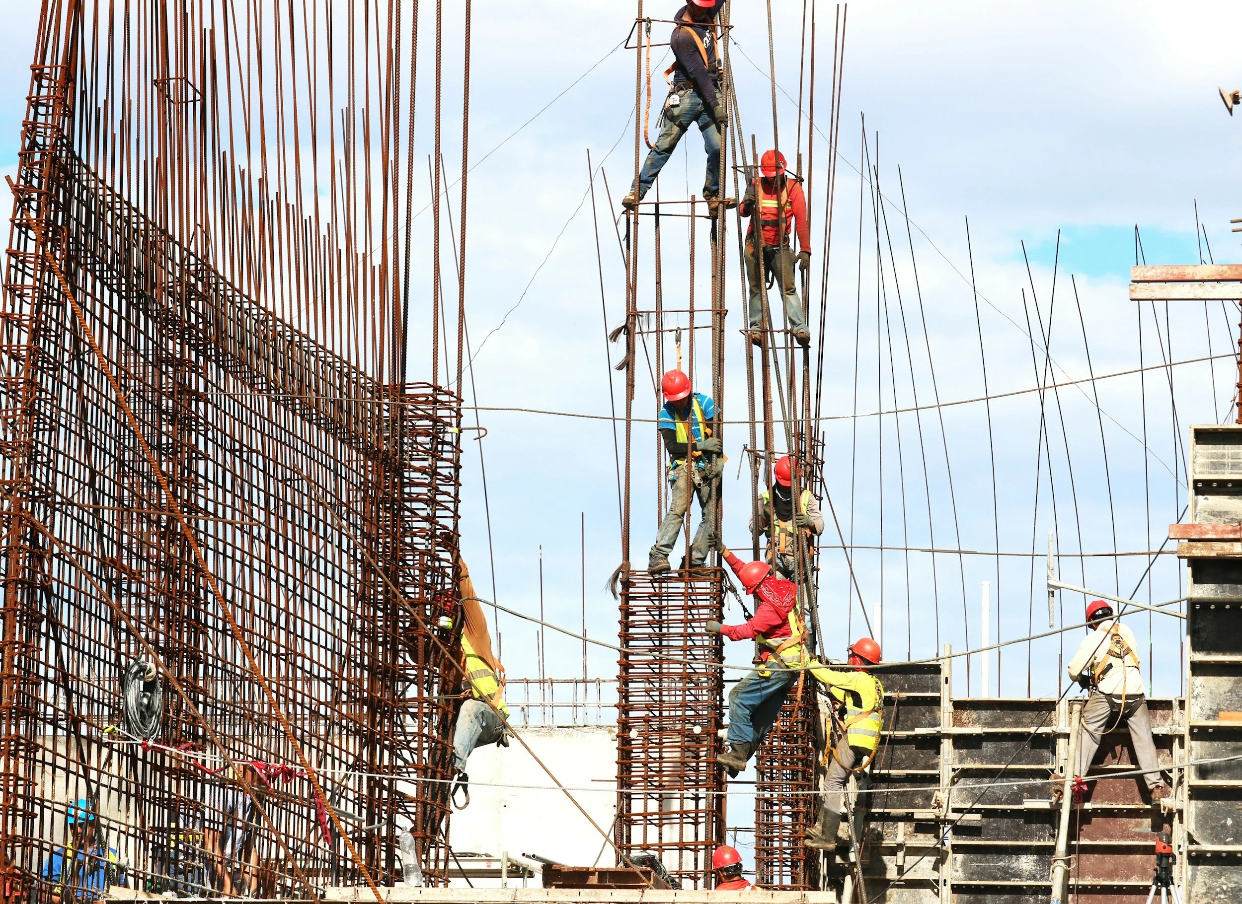 Construction workers wearing helmets and harnesses working on steel rebar framework at a building site.