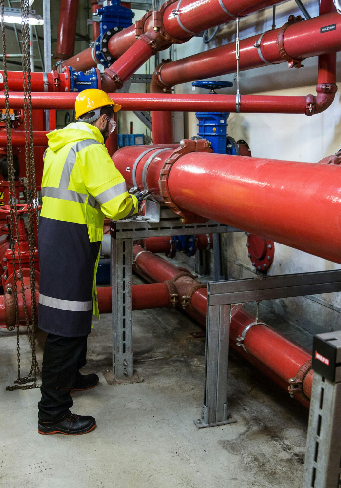 A worker wearing a yellow safety helmet and high-visibility jacket inspecting red industrial pipes in a mechanical room.