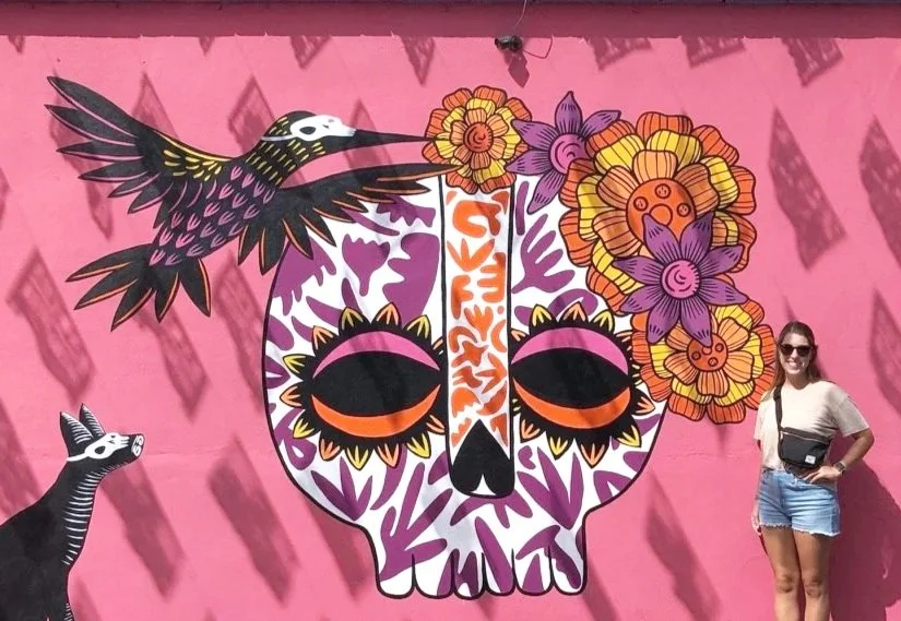 Colorful mural on pink wall featuring a skull decorated with floral motifs, a bird flying above, a striped cat on the left, and a woman in sunglasses and casual clothes standing nearby.