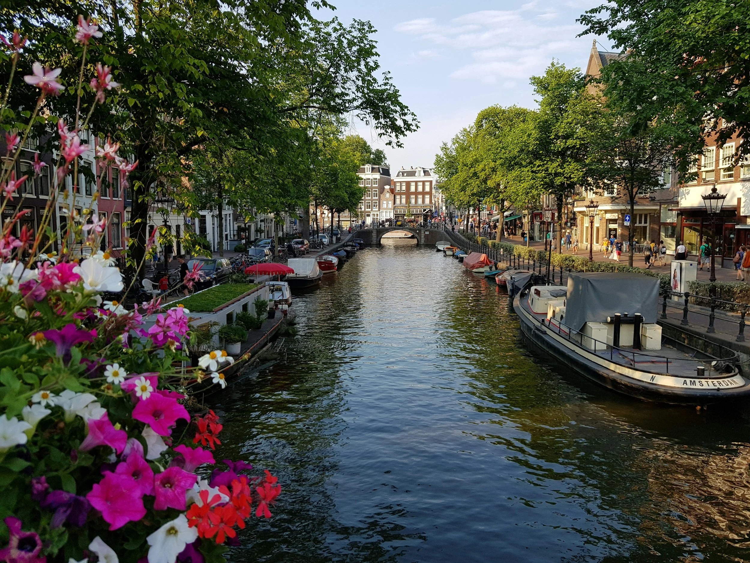 A canal in Amsterdam with boats on the water, a stone bridge in the background, and colorful flowers in the foreground. Trees line the sides of the canal, and buildings with shops and pedestrians are visible along the street.