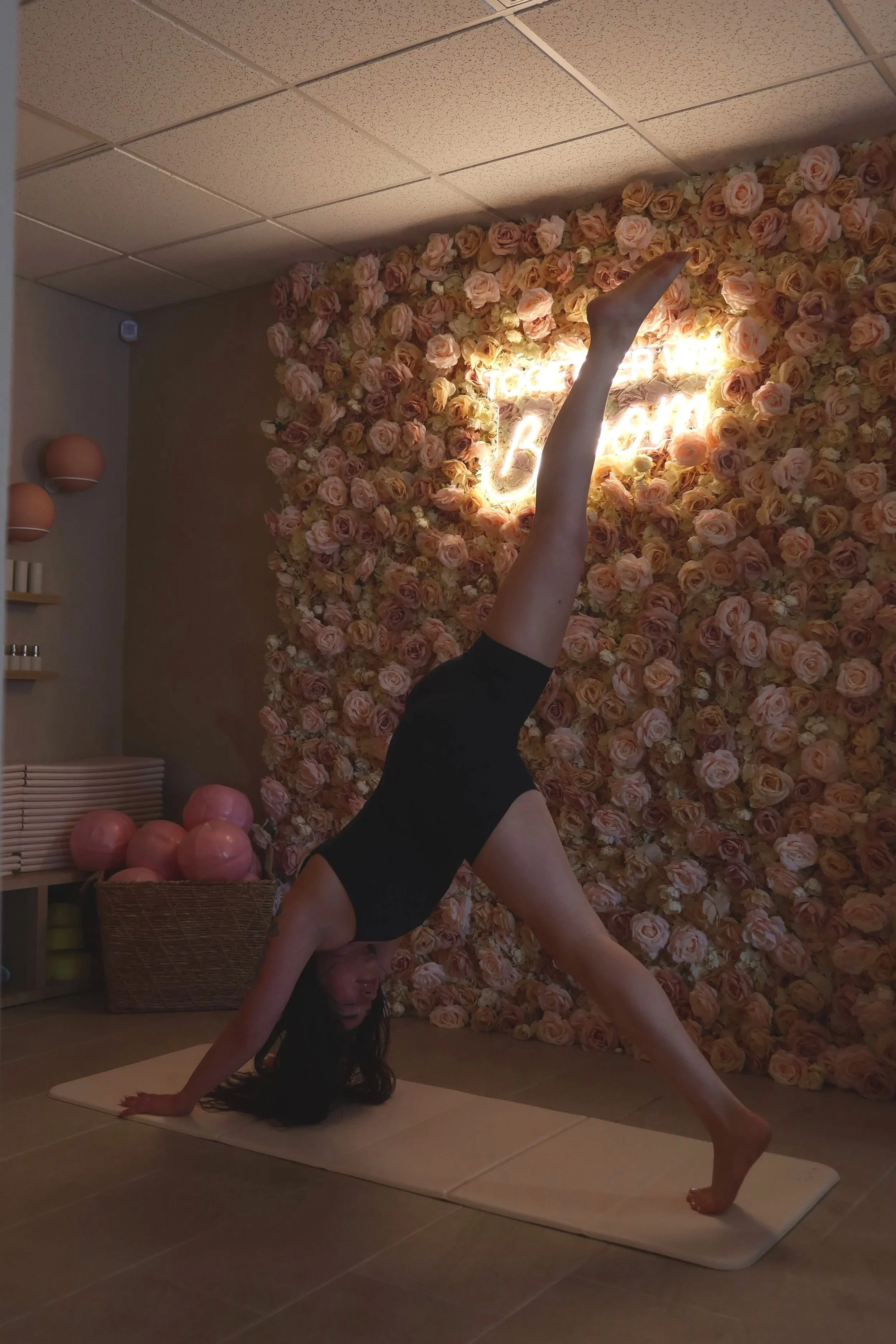 A woman practicing yoga in a handstand pose on a mat in a room with a pink floral wall and neon sign reading 'love be kind'.