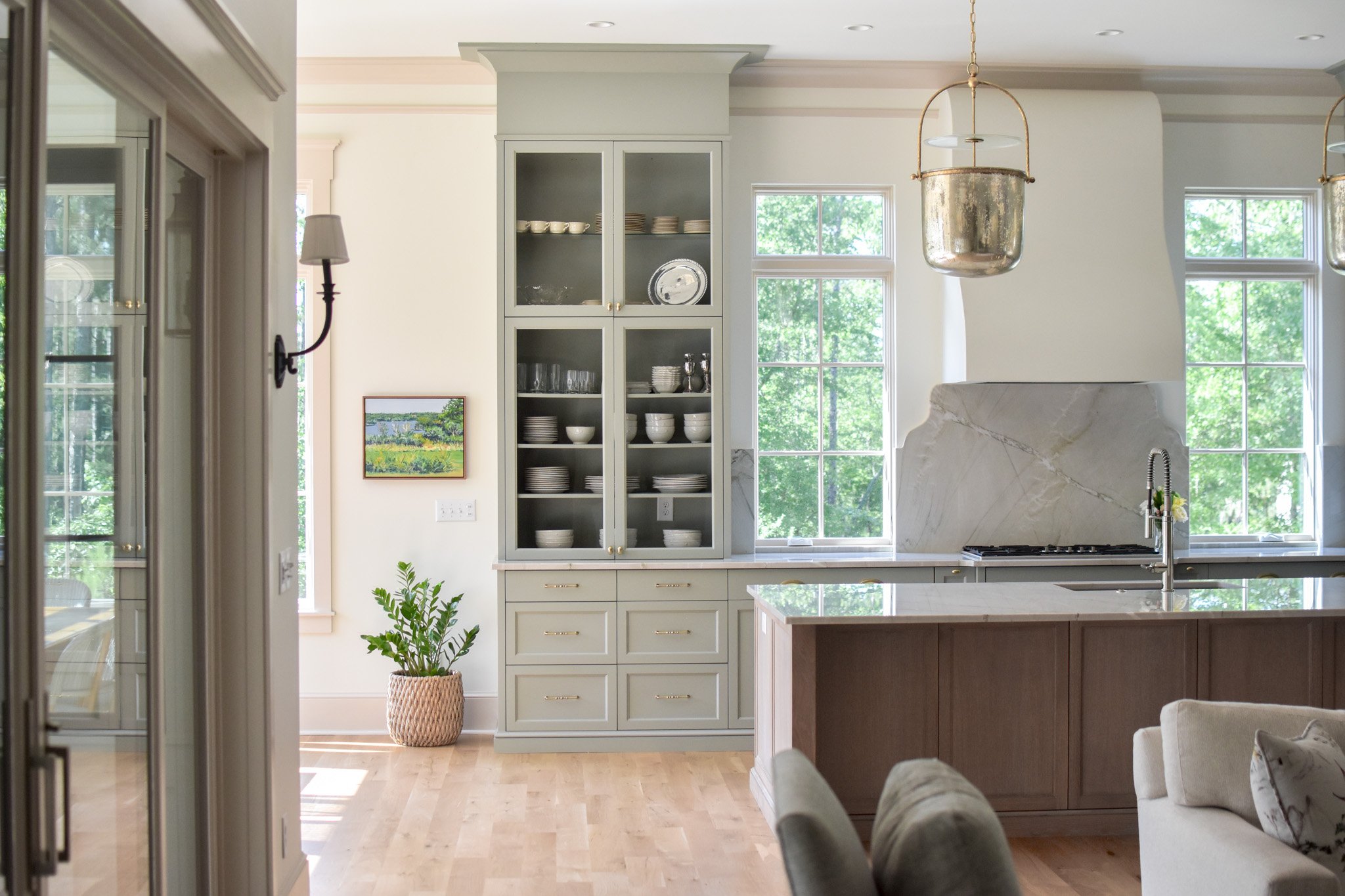 Bright kitchen with white cabinets, open glass-front display cabinet, large windows with green trees outside, kitchen island, marble backsplash, hanging pendant lights, and a potted plant near the wall.
