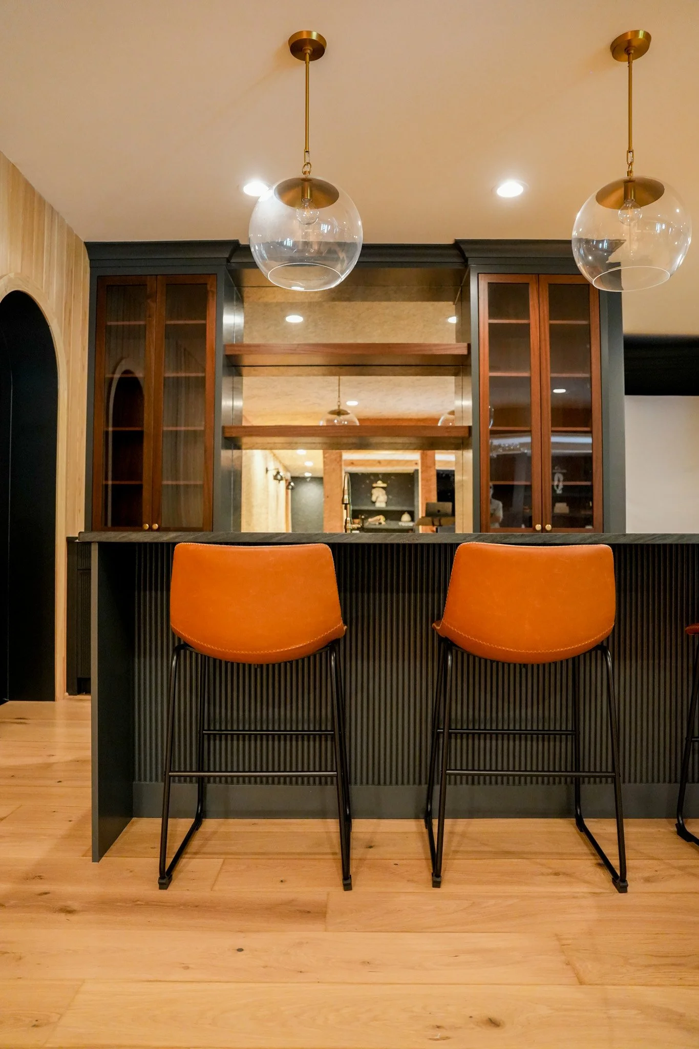 Interior view of a kitchen or bar area with two orange bar stools, a black and wooden counter, glass globe pendant lights, and wooden cabinetry.