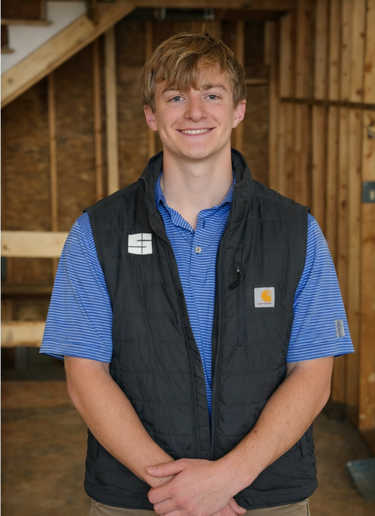 A young man with orange hair smiling, wearing a blue striped polo shirt and a black Carhartt vest, standing indoors in a wooden structure.
