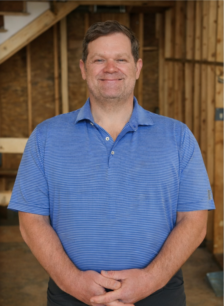 A smiling man in a blue striped polo shirt standing inside a wooden structure, possibly a workshop or garage.