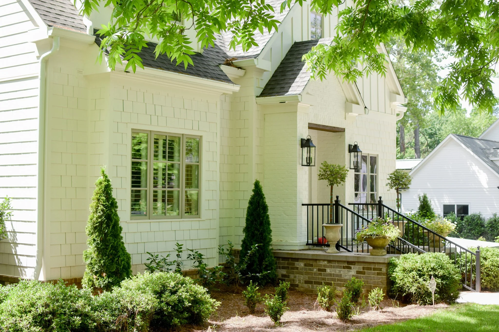 Front yard of a cream-colored house with a small porch, black railings, potted plants, tall trees, and trimmed bushes.