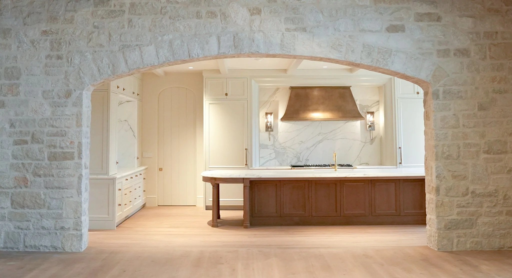Kitchen viewed through a large stone archway, featuring white cabinets, a marble backsplash, and a wooden kitchen island with a marble countertop.