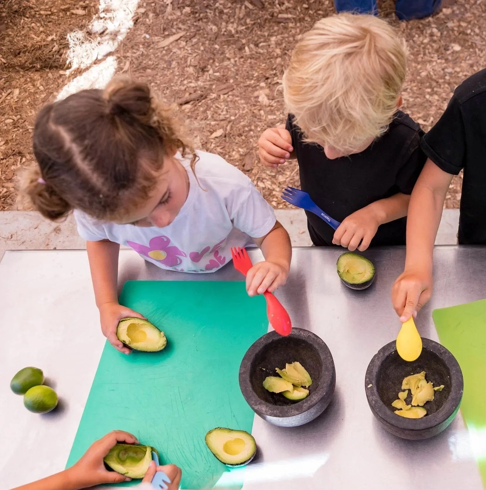 Two children are preparing avocados at a table, scooping out their flesh with colorful forks.