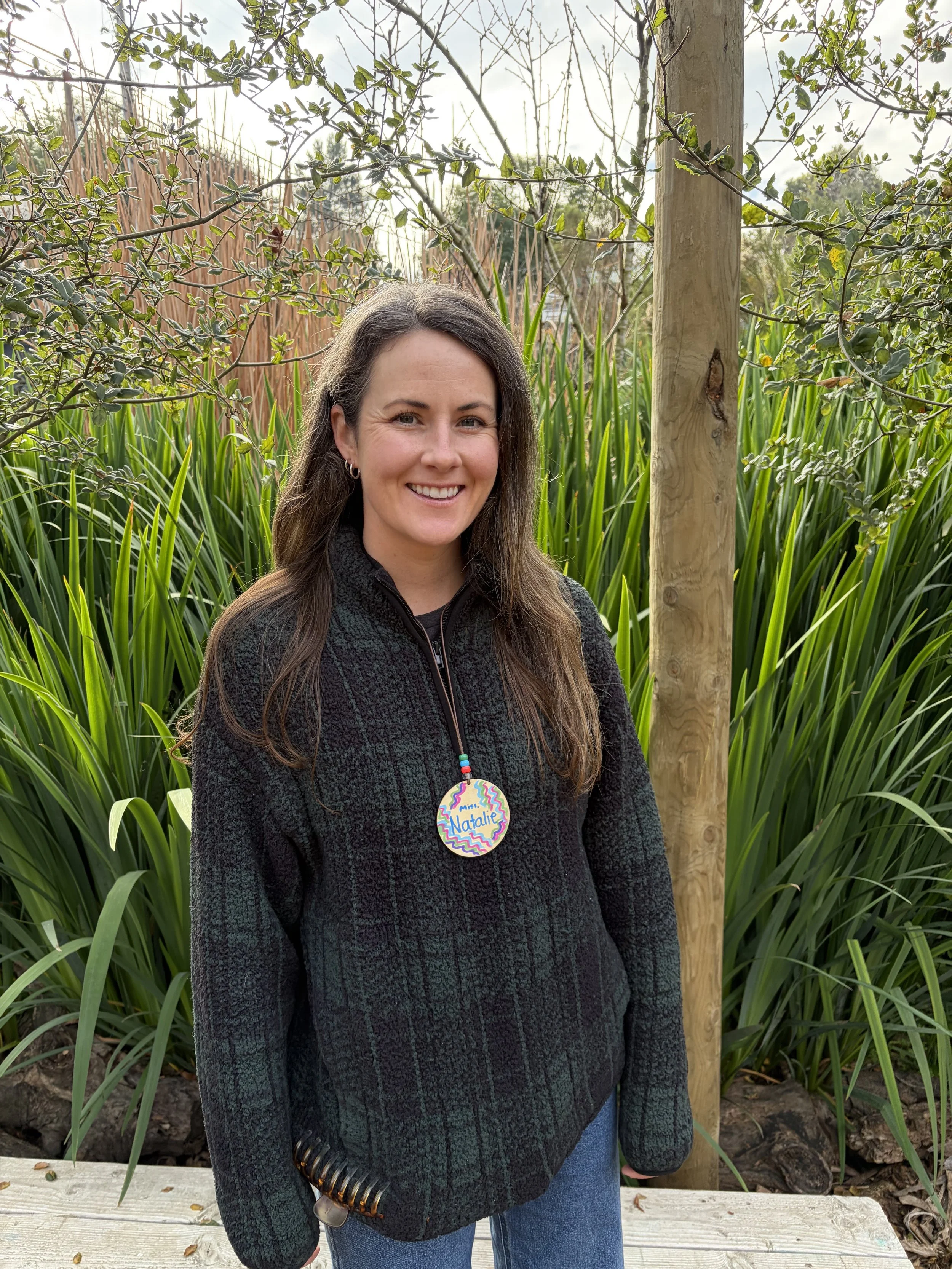 A woman outdoors in a garden with green plants and trees, smiling at the camera, wearing a black jacket and a birthday badge that says 'Happy Birthday Natalie'.