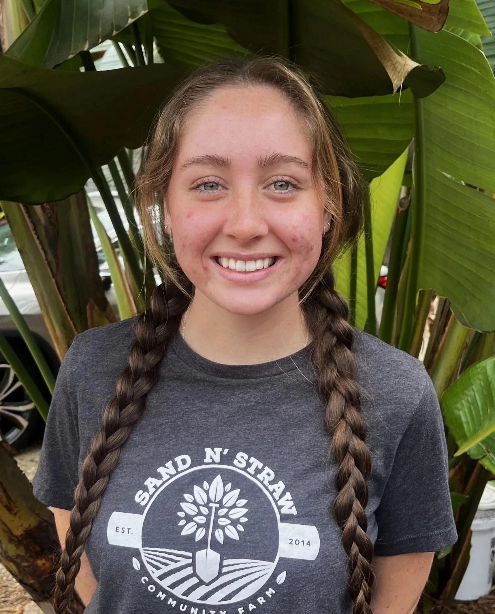 Smiling young woman with long braided hair wearing a gray t-shirt with a logo for Sand N' Straw Community Farm, standing in front of large green plants.