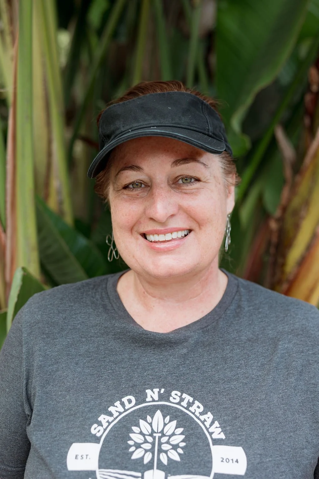 A smiling woman with short hair, wearing a black cap, earrings, and a dark grey t-shirt with a logo reading 'Sand N' Street,' standing outdoors in front of large green leaves.