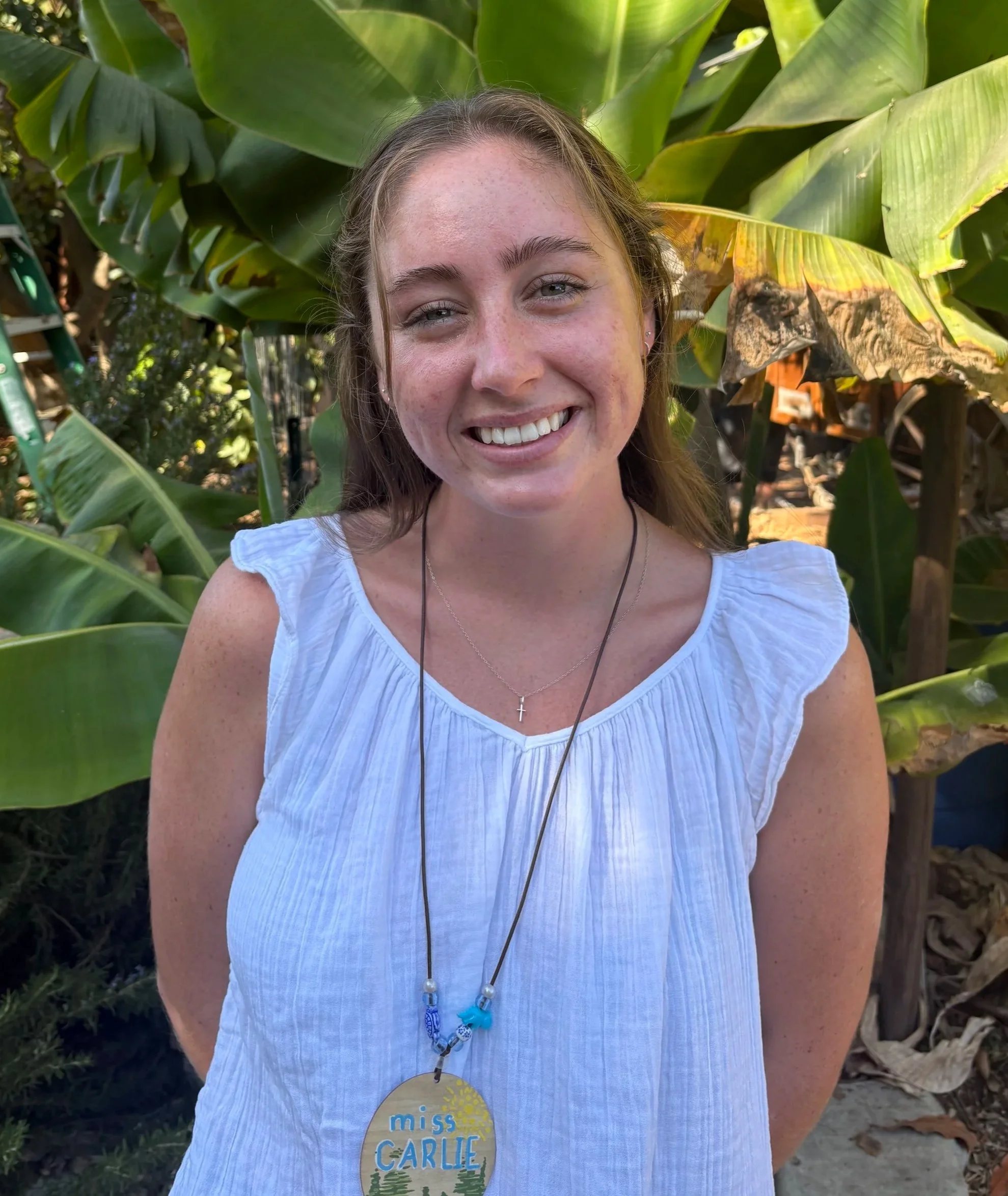 Smiling young woman with long braided hair wearing a gray t-shirt with a logo for Sand N' Straw Community Farm, standing in front of large green plants.