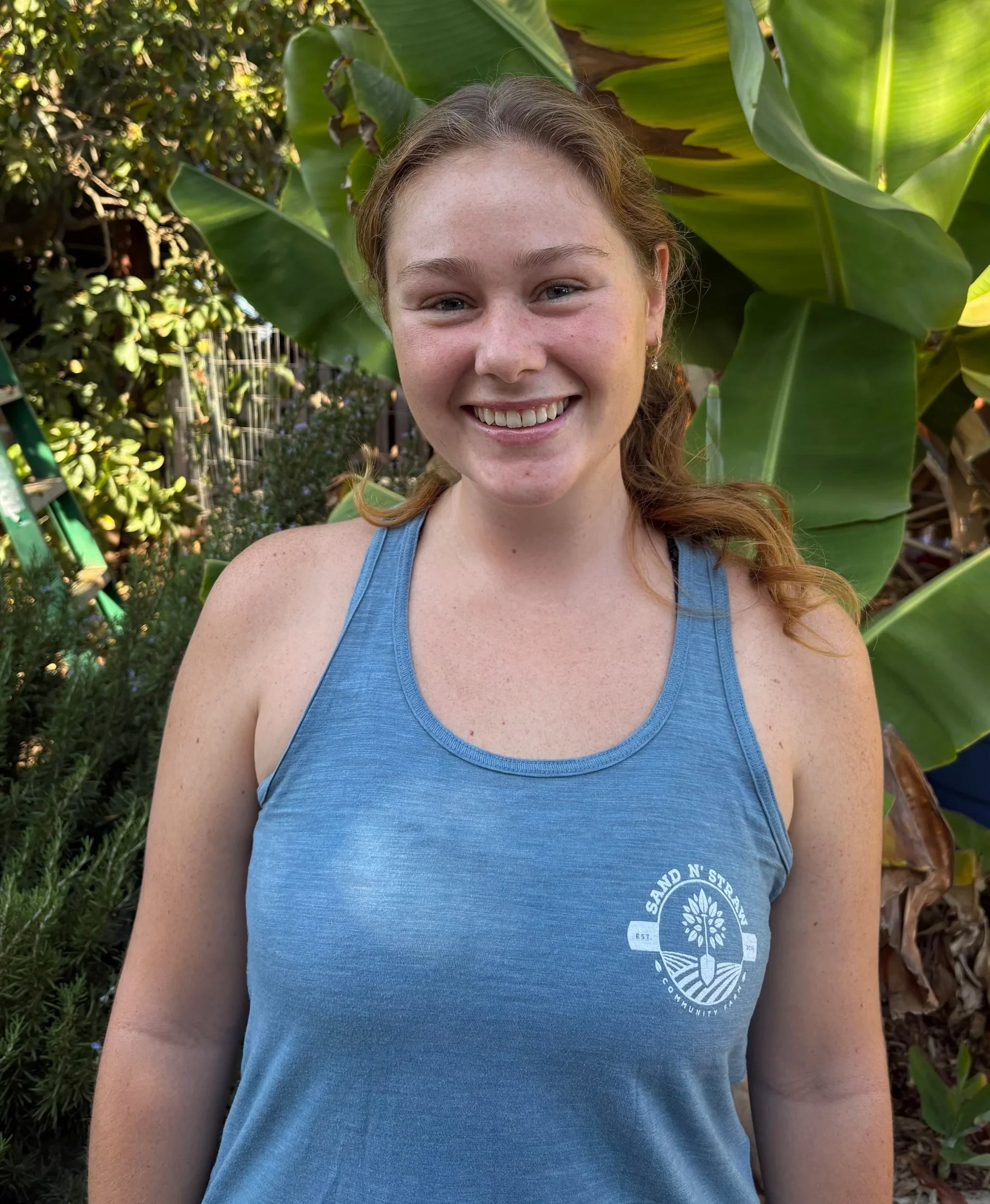 Close-up of a young woman with red hair, blue eyes, and freckles smiling outdoors, wearing a white t-shirt with a Rip Curl logo.
