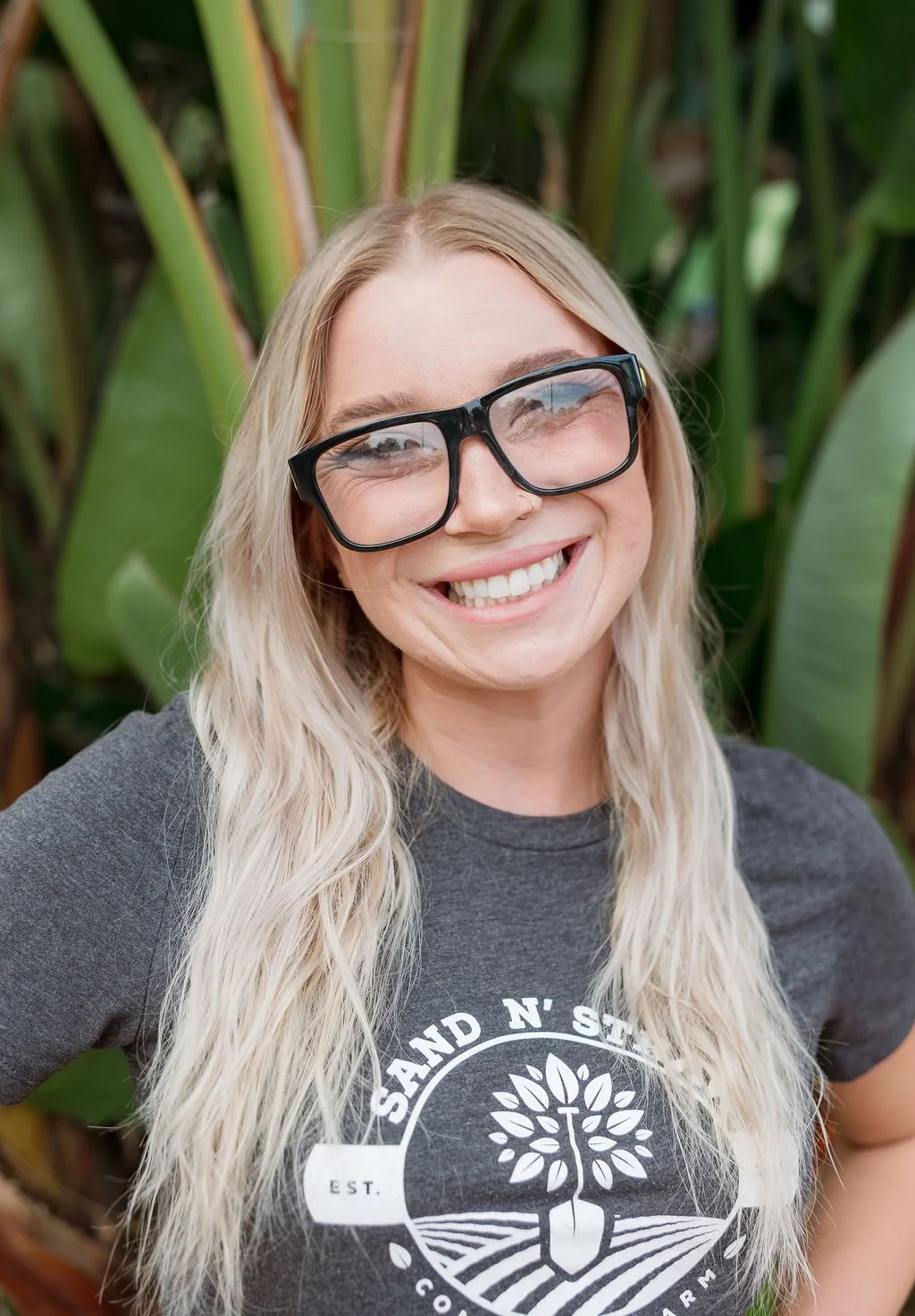 A young woman with long blonde hair, smiling, wearing large black glasses and a dark gray t-shirt with a logo featuring a shovel and the text "Sand N' Soil." She is outdoors, in front of a backdrop of green foliage.