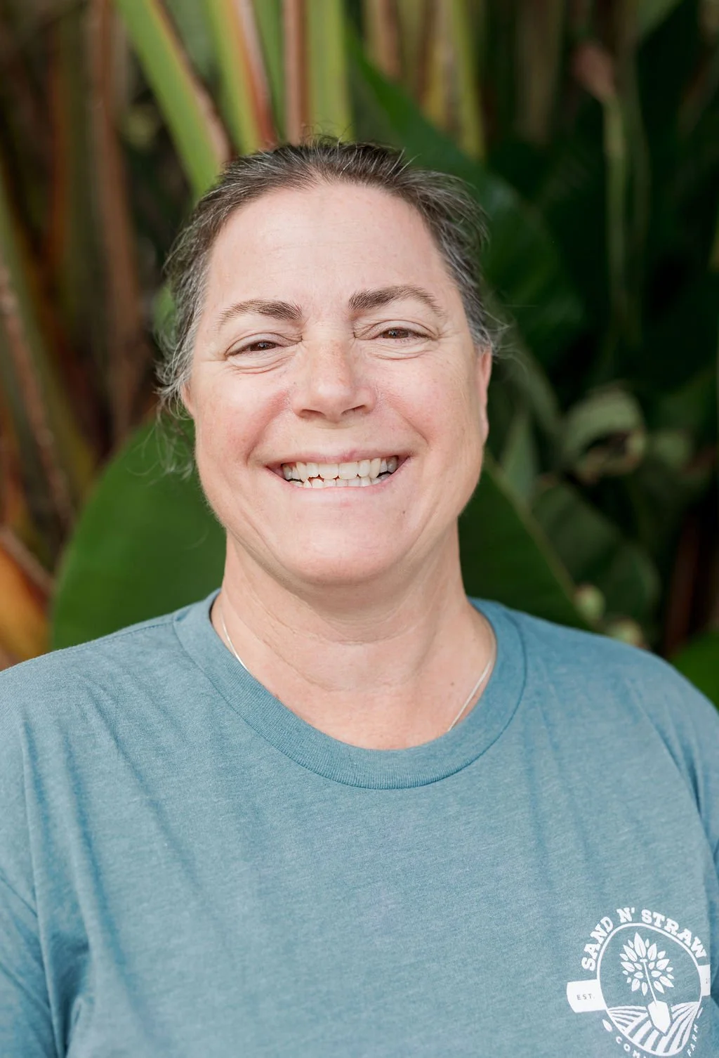 Smiling woman with short gray hair wearing a blue T-shirt with a logo, standing outdoors with green foliage in the background.