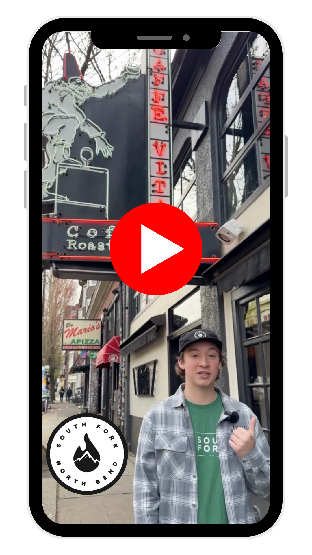 Young man standing outside a restaurant on a city sidewalk, wearing a black cap and a plaid shirt, with a logo that reads 'South Fork North Bend'. The restaurant signs are visible, including a neon sign and a sign for Mario's Pizza.