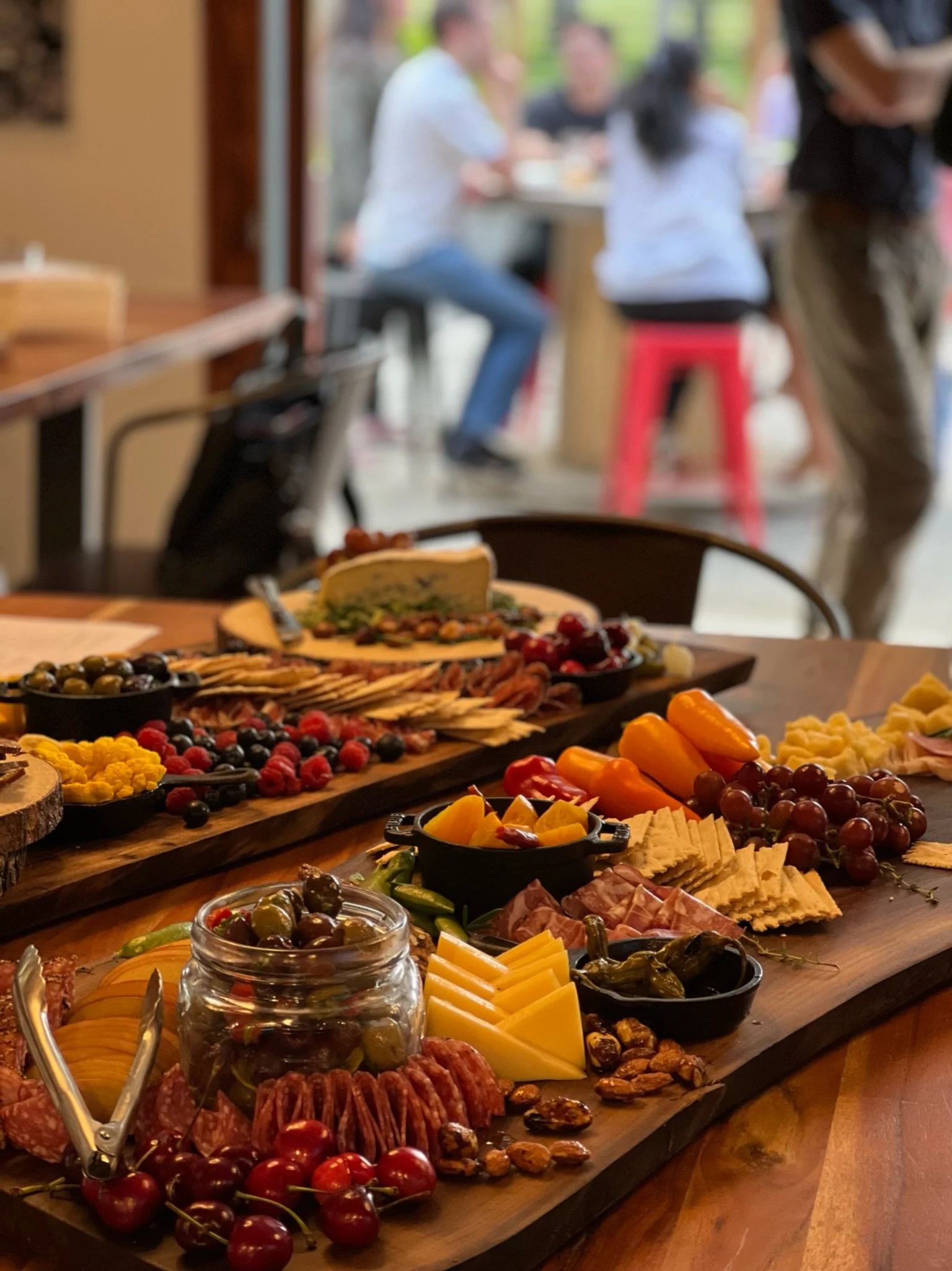 A cheese and charcuterie board with grapes, berries, nuts, sliced cheese, and meats on a wooden table, with a blurred background of people at an outdoor gathering.