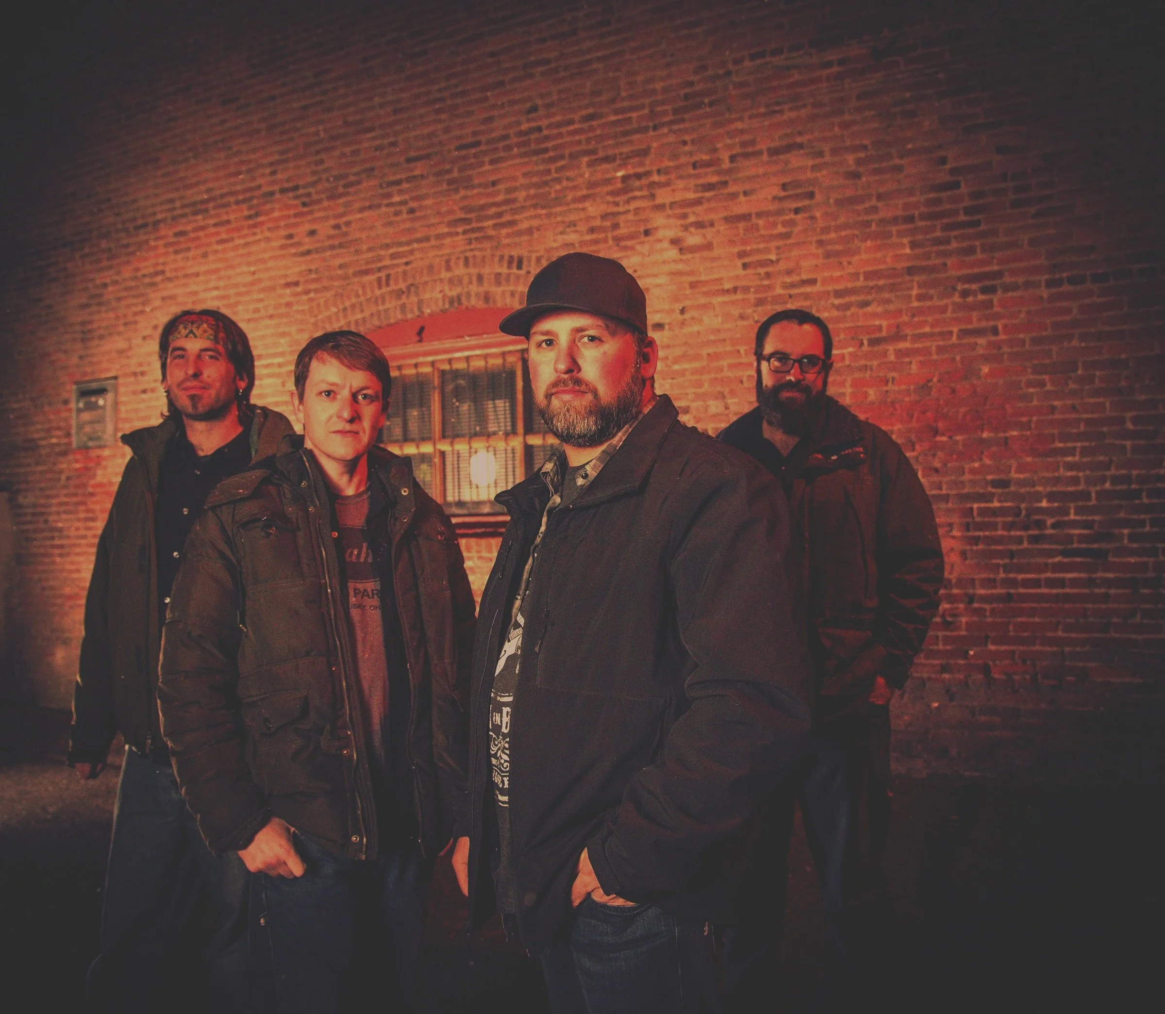 Four men standing in front of a brick wall at night. They are dressed in dark jackets, with the man in the front wearing a cap.