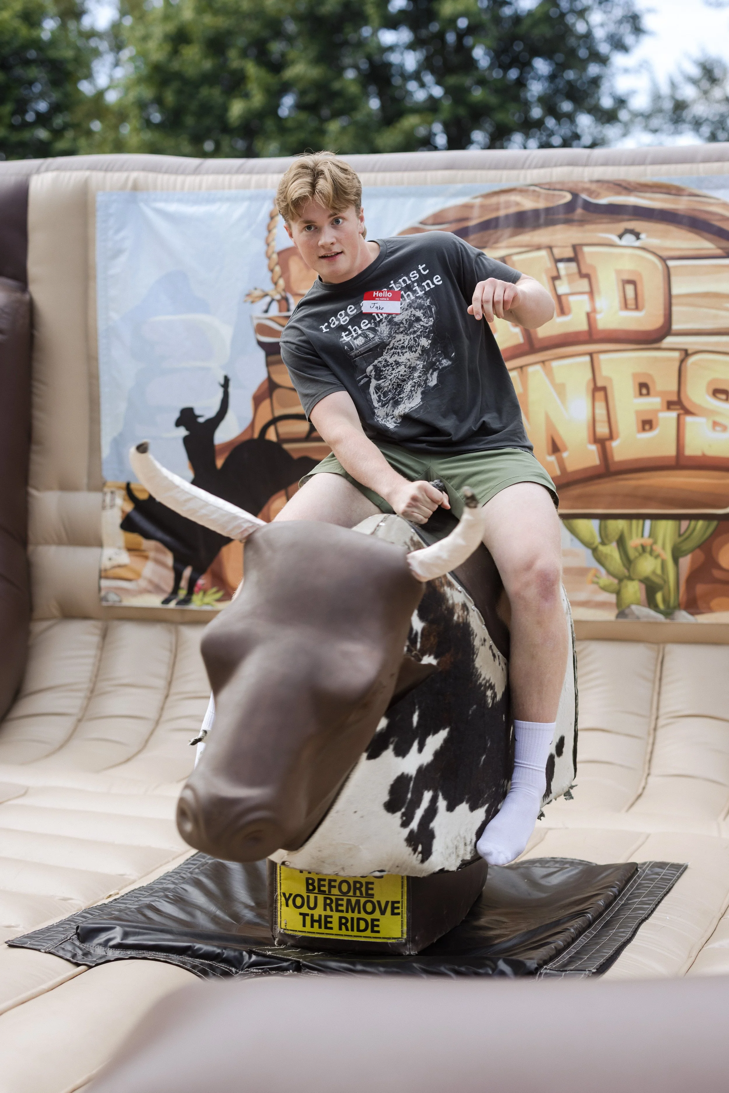 A young man riding a mechanical bull at an outdoor fair or event. He is wearing a dark T-shirt, green shorts, and white socks, with a temporary name tag on his chest. The background features a banner with a Western-themed design. A yellow warning sign on the bull reads, 'BEFORE YOU REMOVE THE RIDE.'