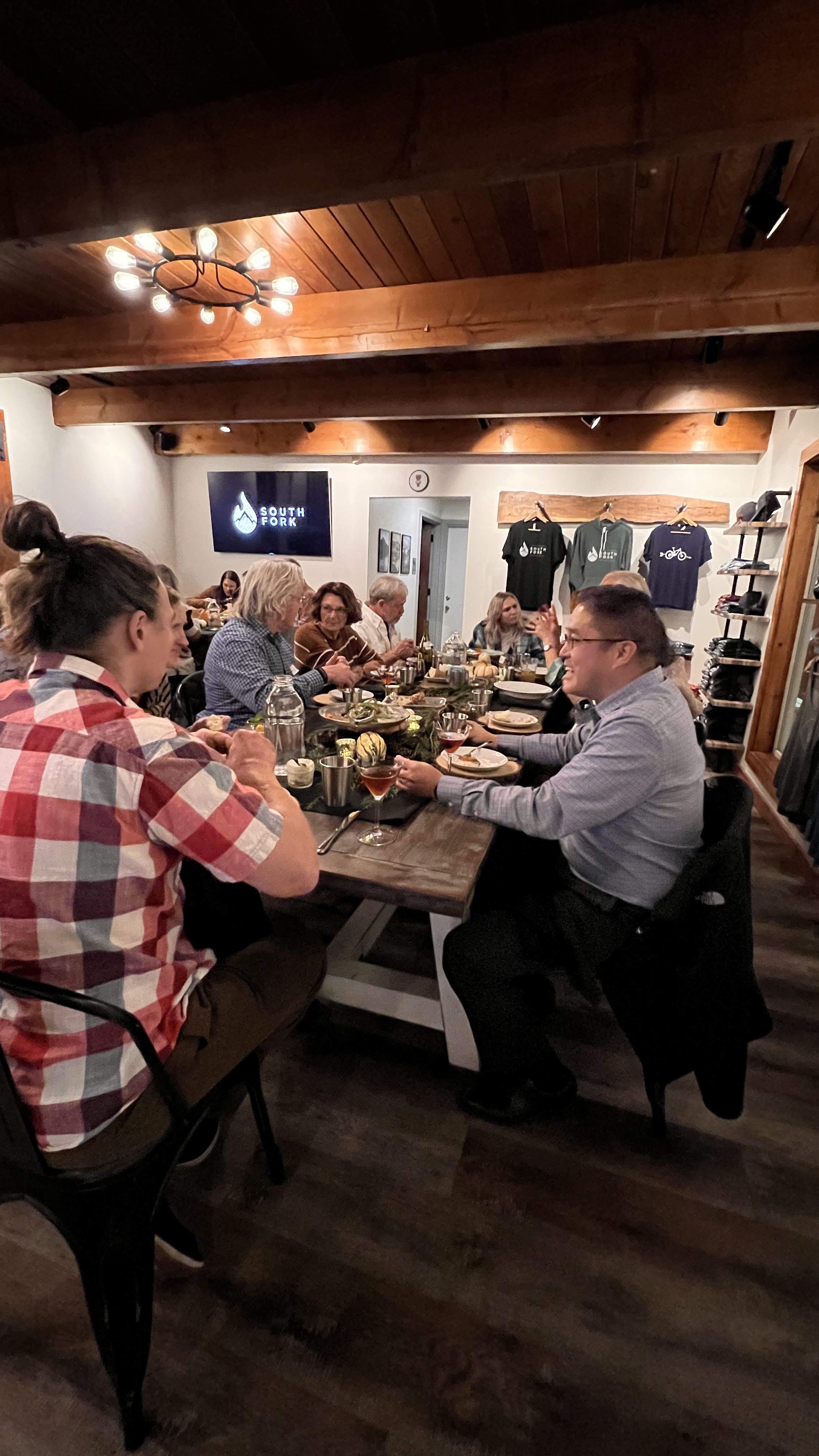 People gathered around a long dining table enjoying a meal in a cozy, rustic restaurant with wood-paneled ceiling and wall art.