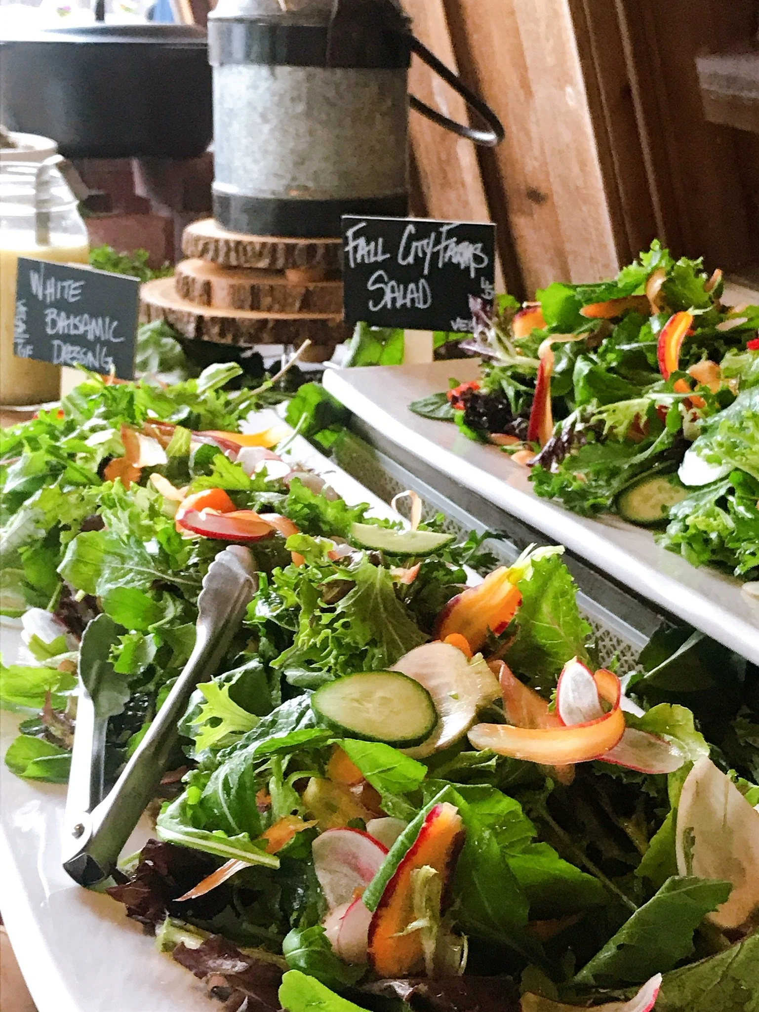 Fresh mixed greens salad with sliced radishes, cucumbers, and carrots, with chalkboard signs indicating different salad options in the background.