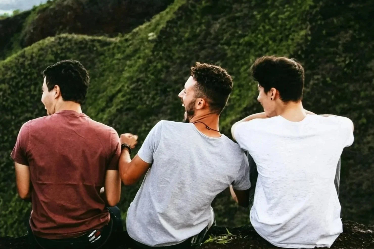 Three young men sitting outdoors with lush green hills in the background, enjoying each other's company and smiling.