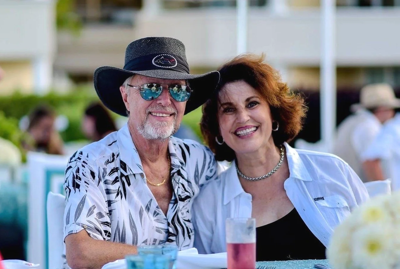 A smiling elderly couple sitting outdoors at a table during a sunny day, with the man wearing sunglasses, a wide-brimmed hat, and a patterned shirt, and the woman in a white blouse and jewelry.