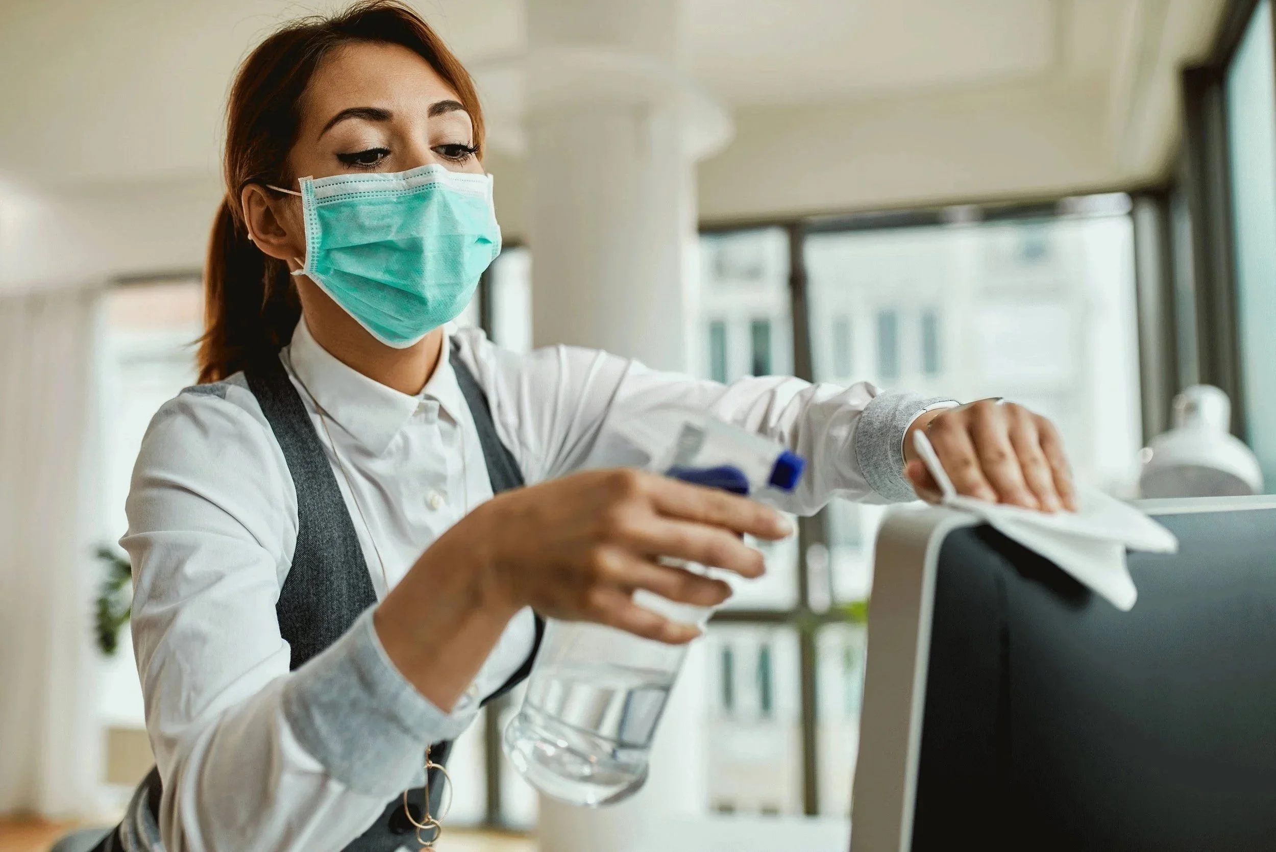 A woman wearing a face mask cleaning a computer monitor with a cloth.