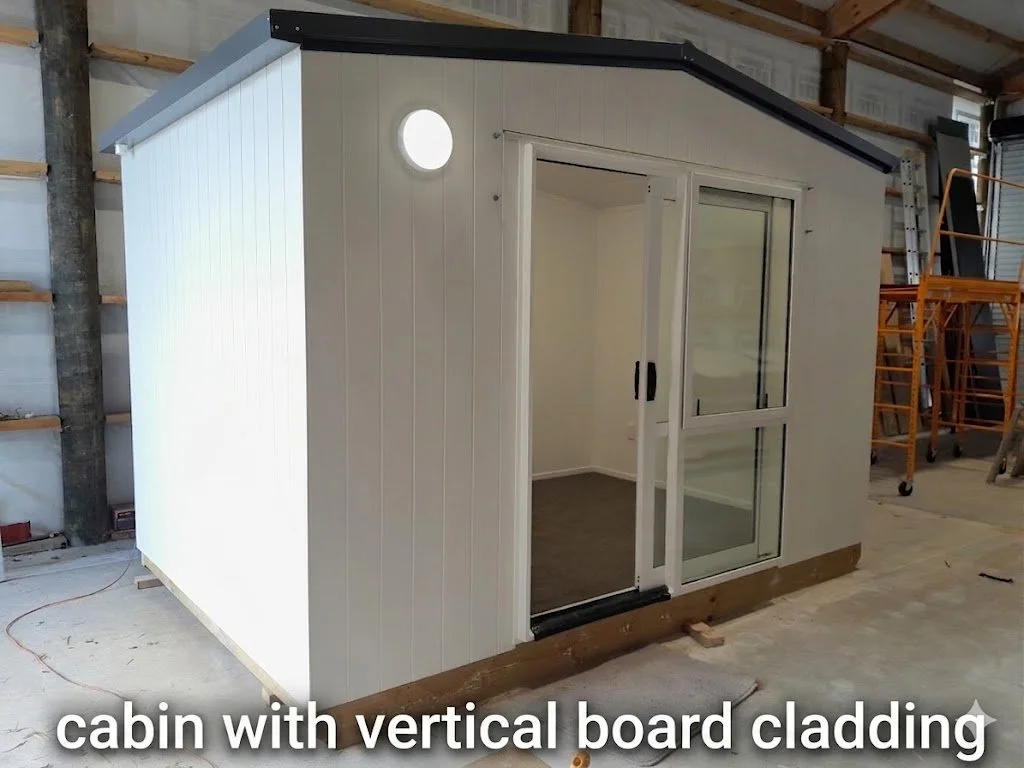 Small cabin with vertical board siding, sliding glass door, circular window, and sloped roof, situated inside a warehouse or garage.