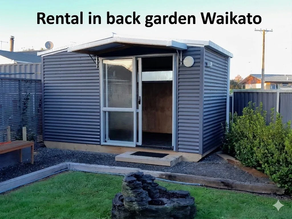 Small backyard garden shed with a sliding glass door, surrounded by a green lawn, plants, and a gravel area.