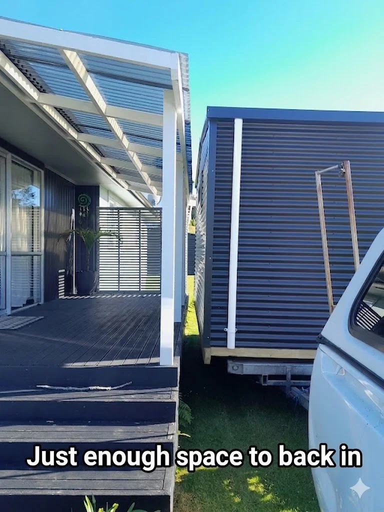 A small deck area with black flooring and a potted plant, next to a narrow space between two structures, with a white truck partially visible on the right and words overlayed that read 'Just enough space to back in'.
