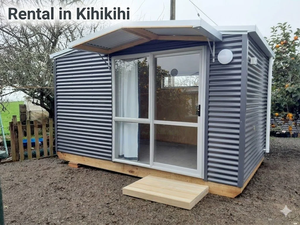 A small, modern tiny home with corrugated metal siding, sliding glass door with curtain, wooden steps, and an overhanging roof, situated outdoors in a yard with trees and orange trees.