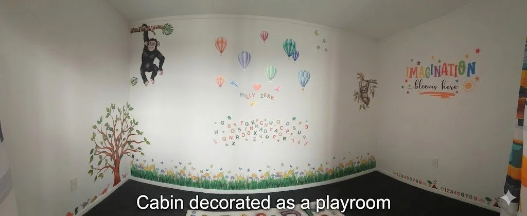 Colorful children’s mural with hot air balloons, a monkey hanging from a branch, a smiling tree, and a rainbow-colored sign that reads "IMAGINATION blooms here" in a cabin playroom.