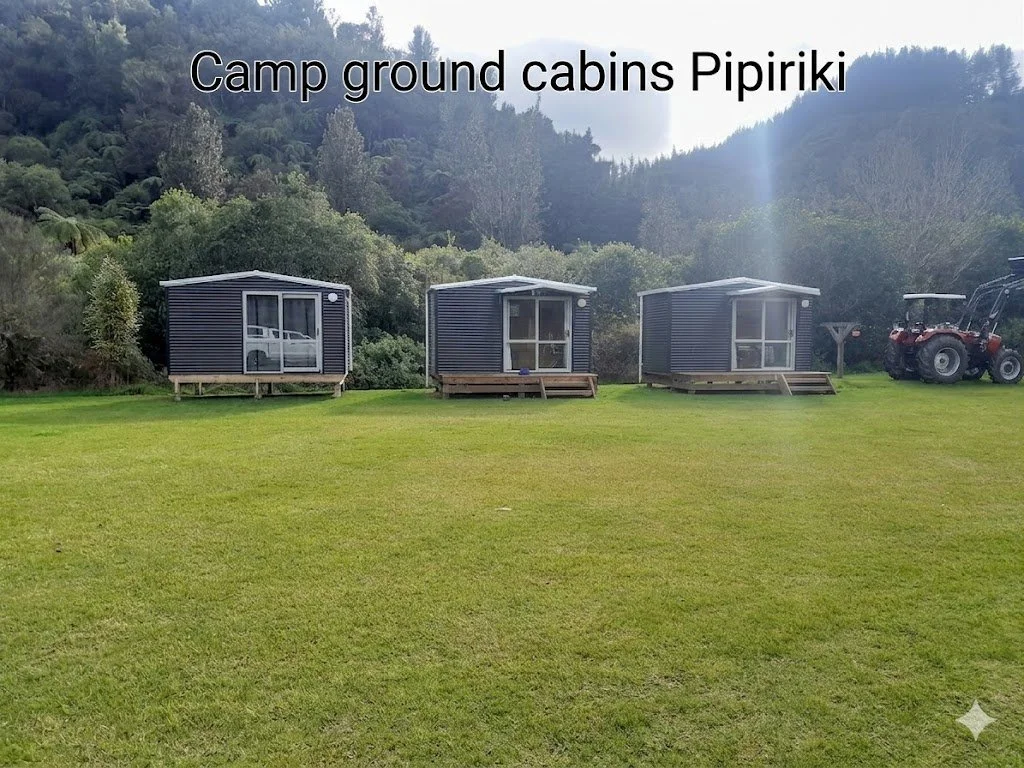 Three small gray cabin structures with stairs are lined up on a grassy field, with a forested hillside in the background and a tractor on the far right. The image has a caption that reads "Camp ground cabins Pipiriki."