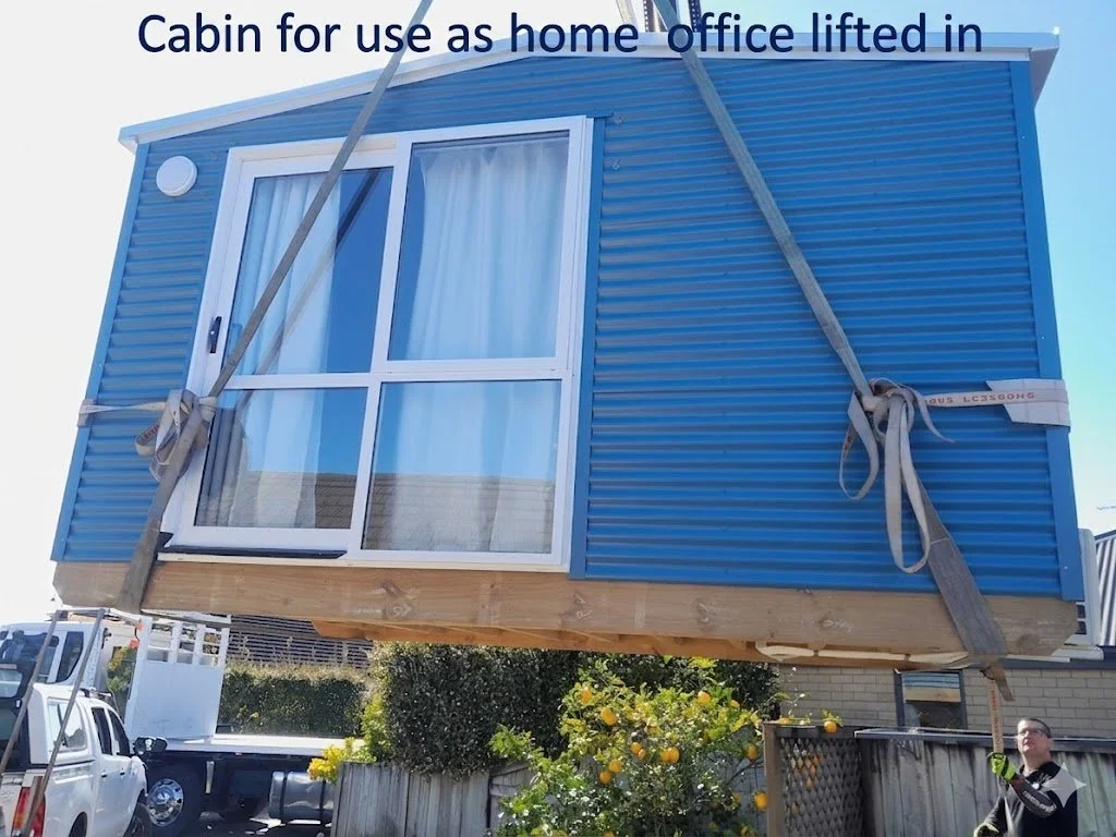A blue tiny home or cabin with large glass windows, lifted in the air using straps and a crane, with a caption 'Cabin for use as home office lifted in'.