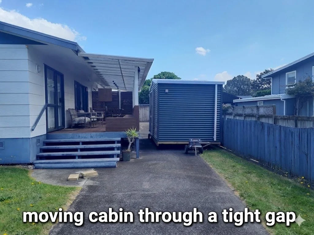 A tiny blue moving cabin being moved through a narrow gap between a house and the fence.