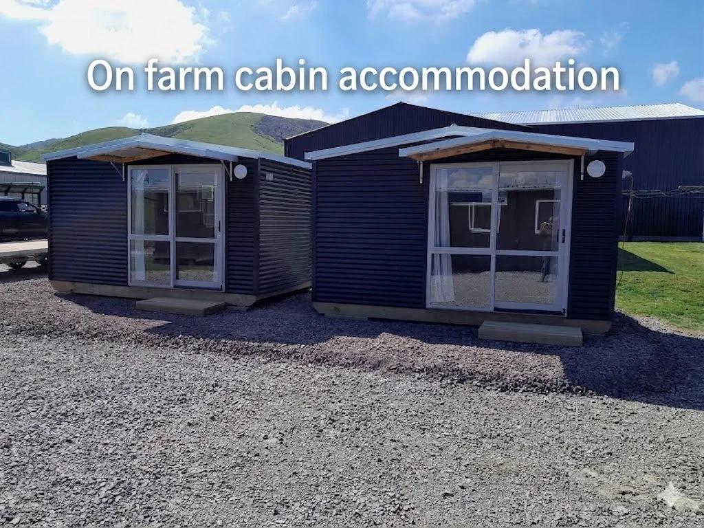 Two small black farm cabins with glass sliding doors, set on gravel ground, under a partly cloudy sky, with green hills in the background.