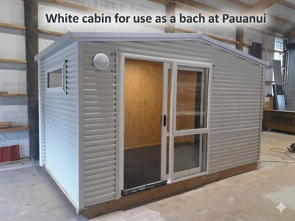 A white metal cabin with a sliding glass door, small window, and round vent, inside a warehouse, labeled as a bach for use at Pauanu.